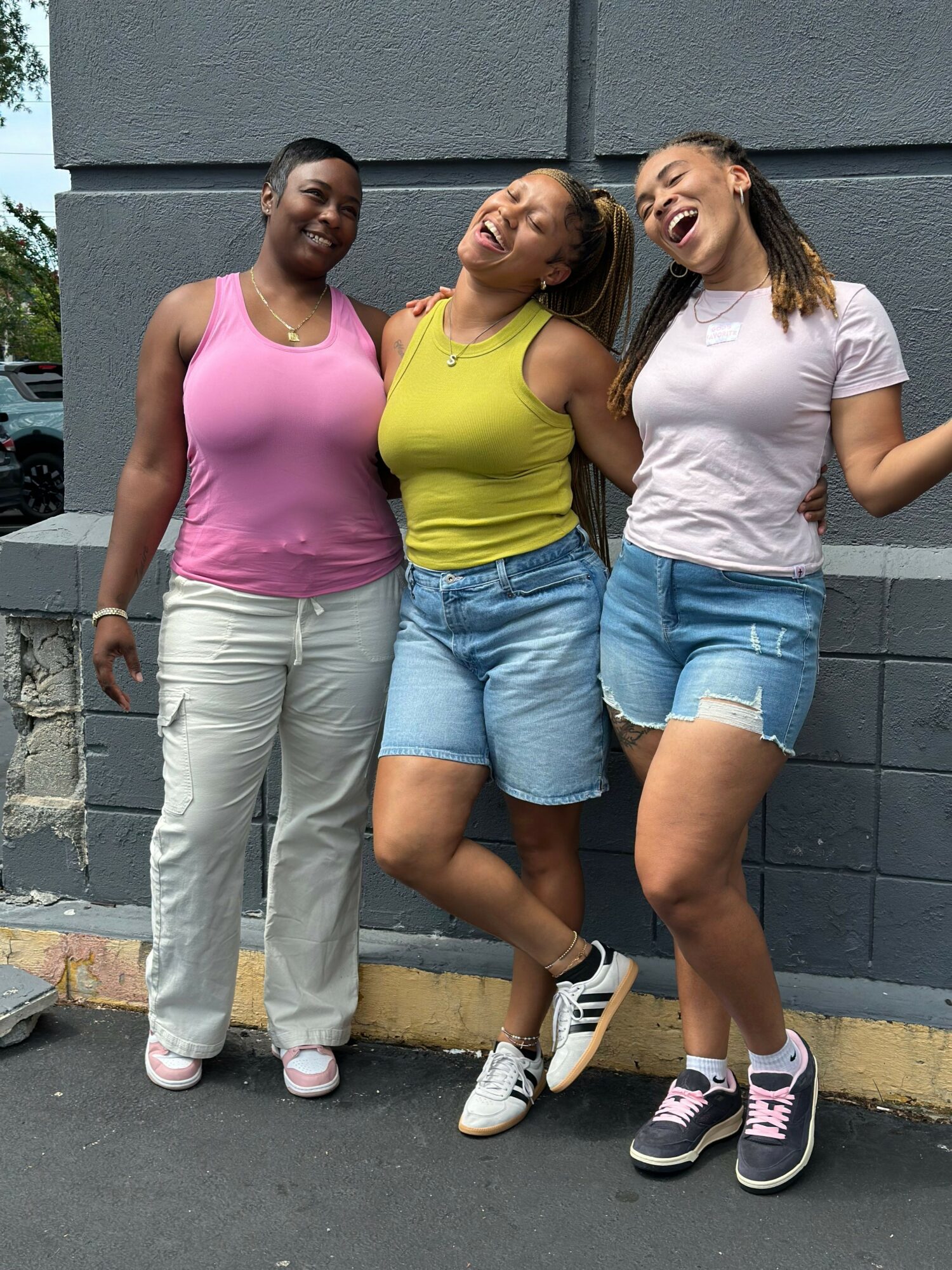 Three women standing outdoors against a gray wall, smiling, wearing casual summer clothes and sneakers.