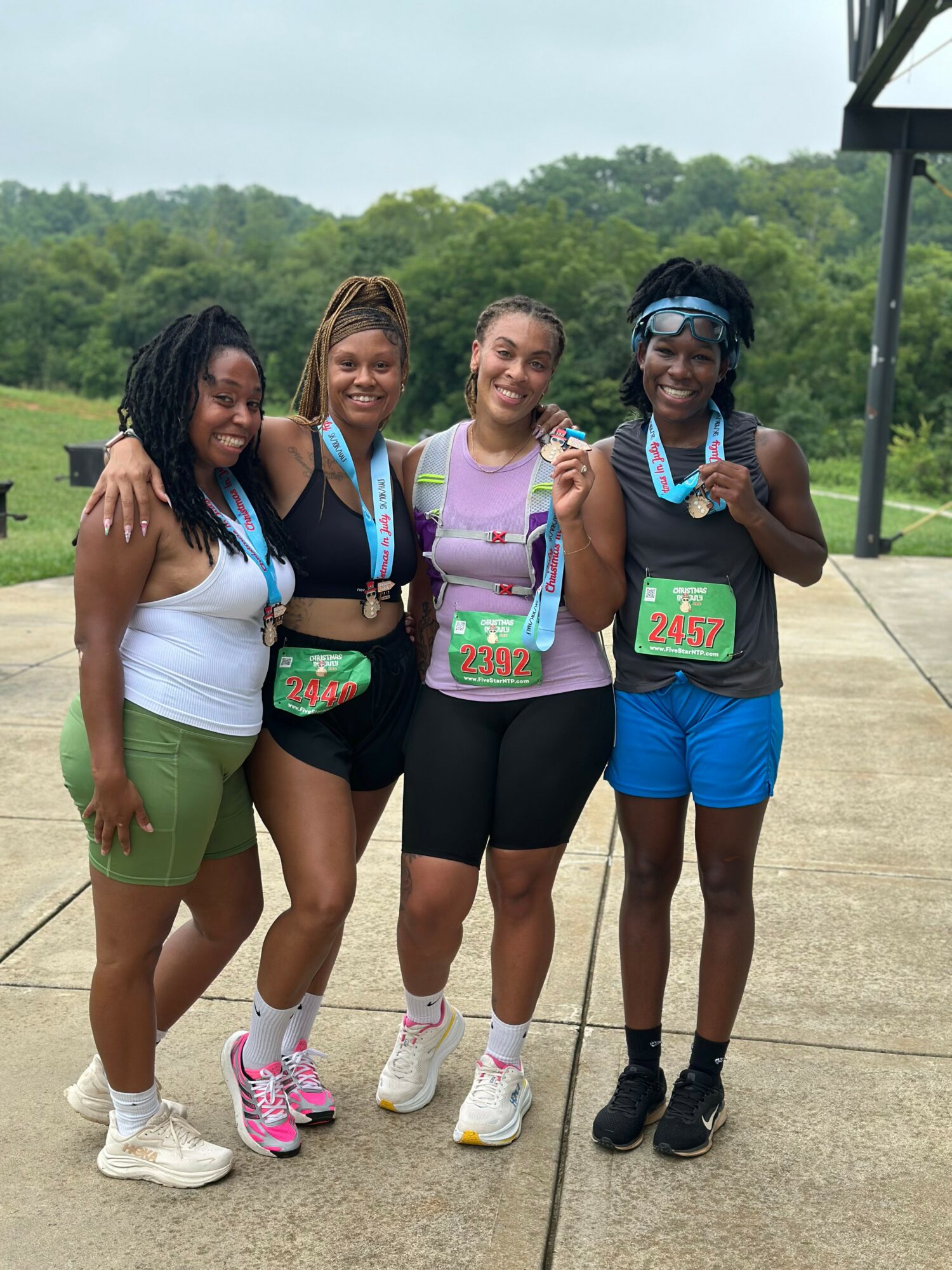 Four women standing outdoors, smiling, wearing athletic clothing and medals, with trees and a cloudy sky in the background.