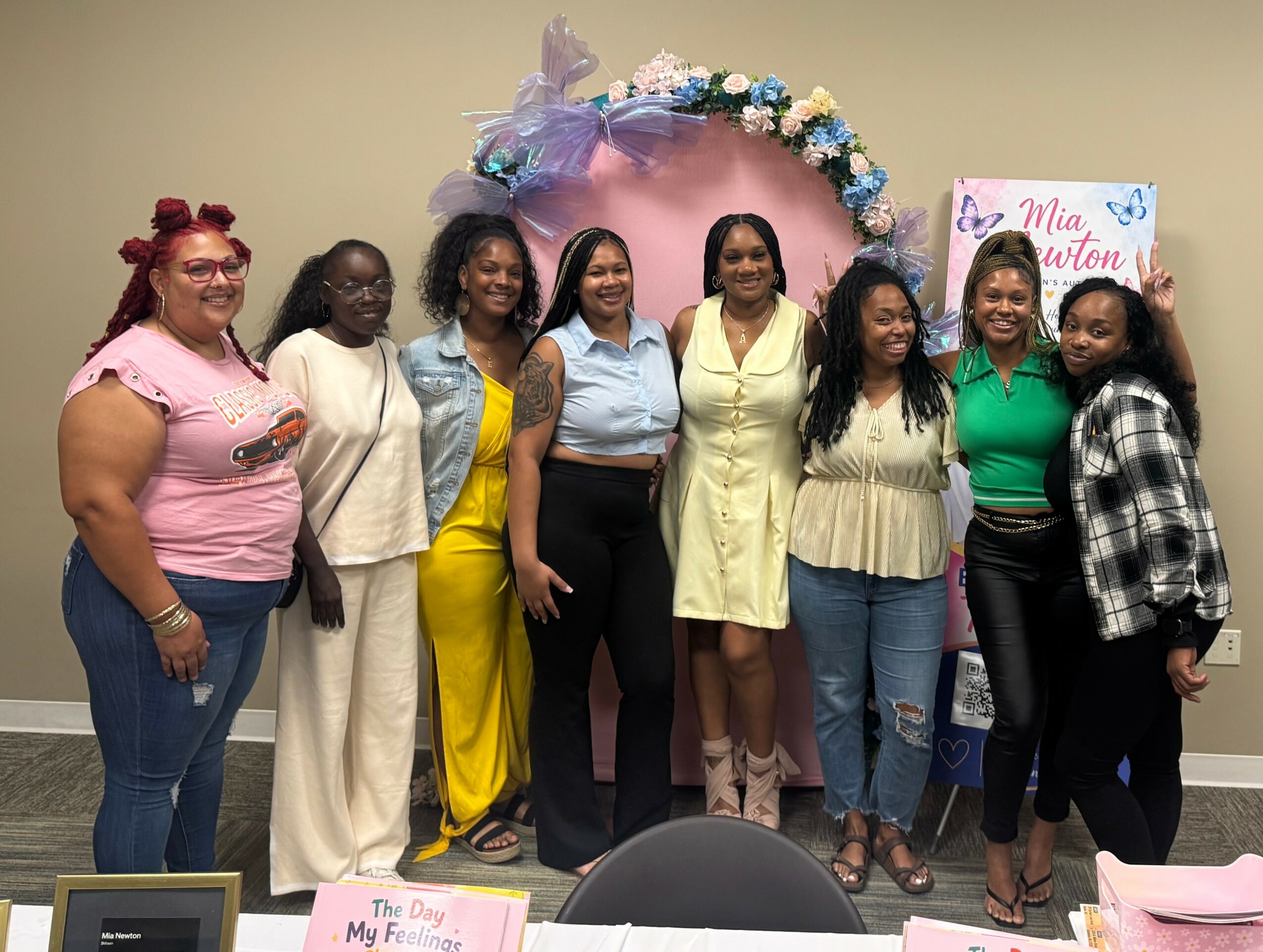 Group of eight women standing in front of a pink and purple floral backdrop, smiling, indoors.