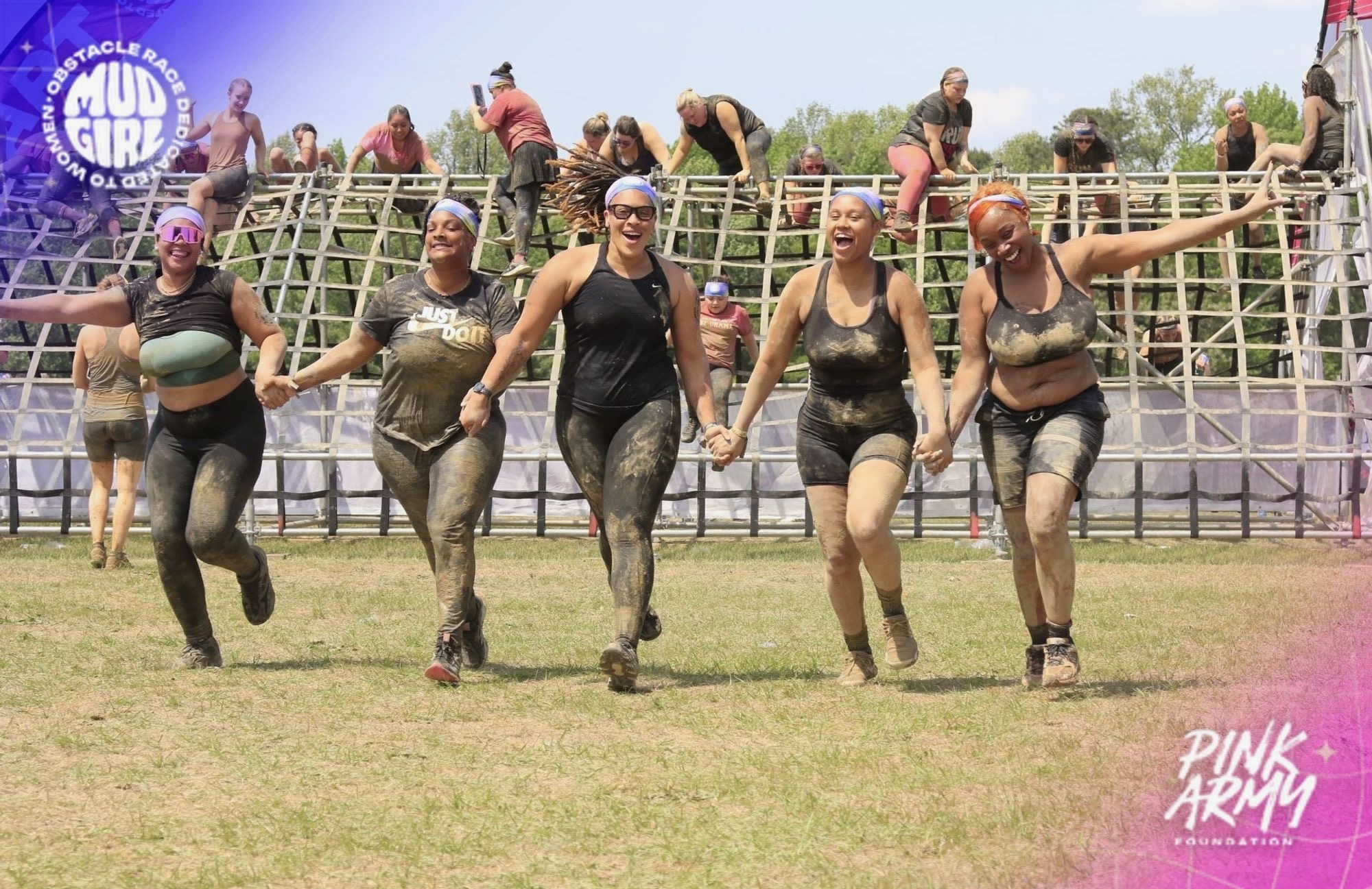 Six women holding hands and running on grass during an outdoor event, with a crowd and metal barriers in background.