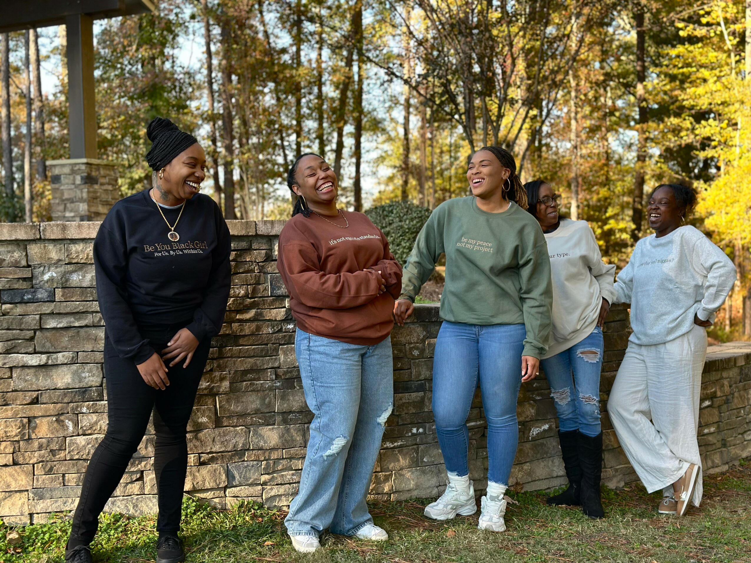Five women stand outdoors, smiling and talking, with trees and a stone wall in the background.