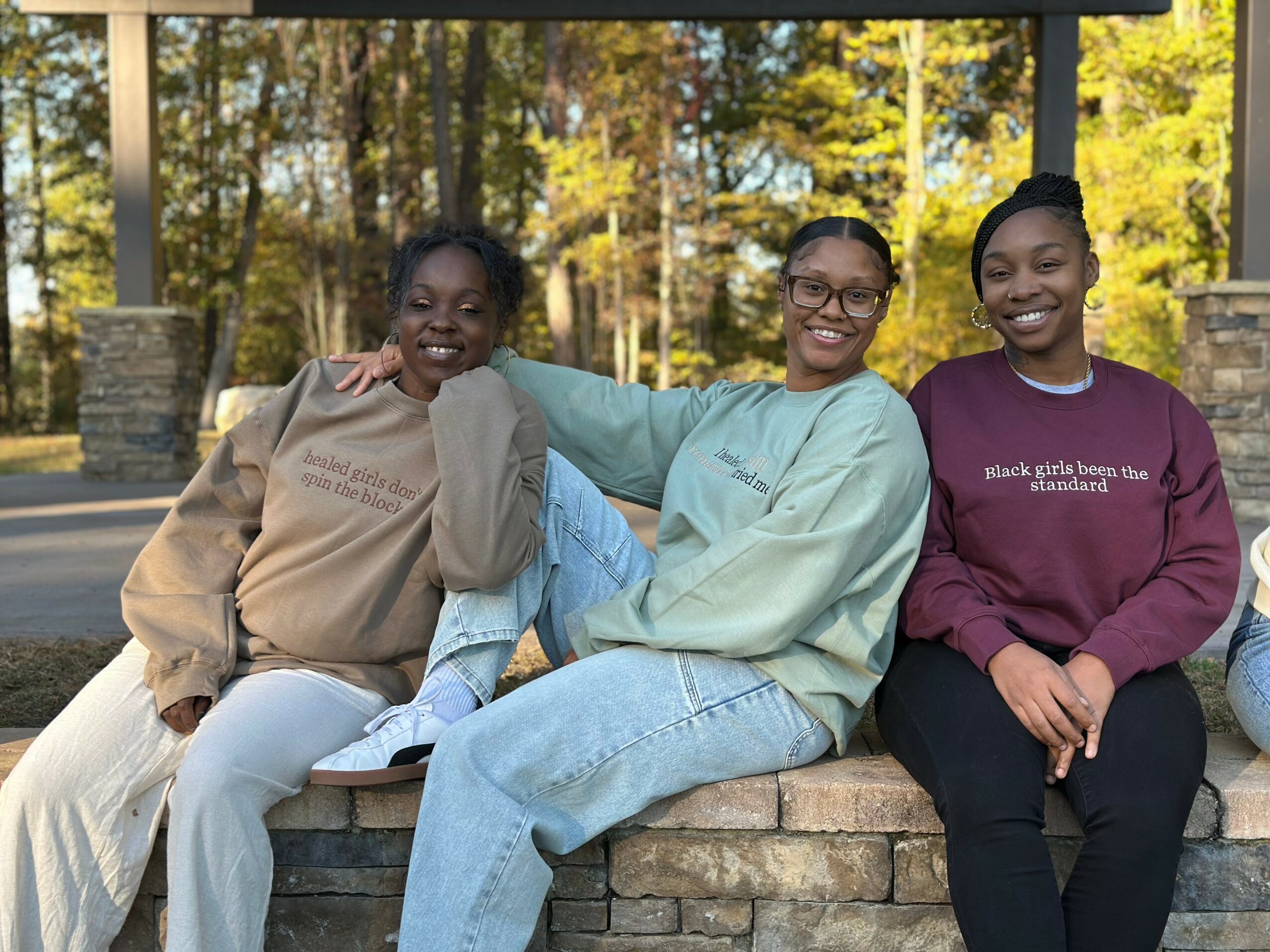 Three smiling women sitting on a stone bench outdoors with trees in the background.
