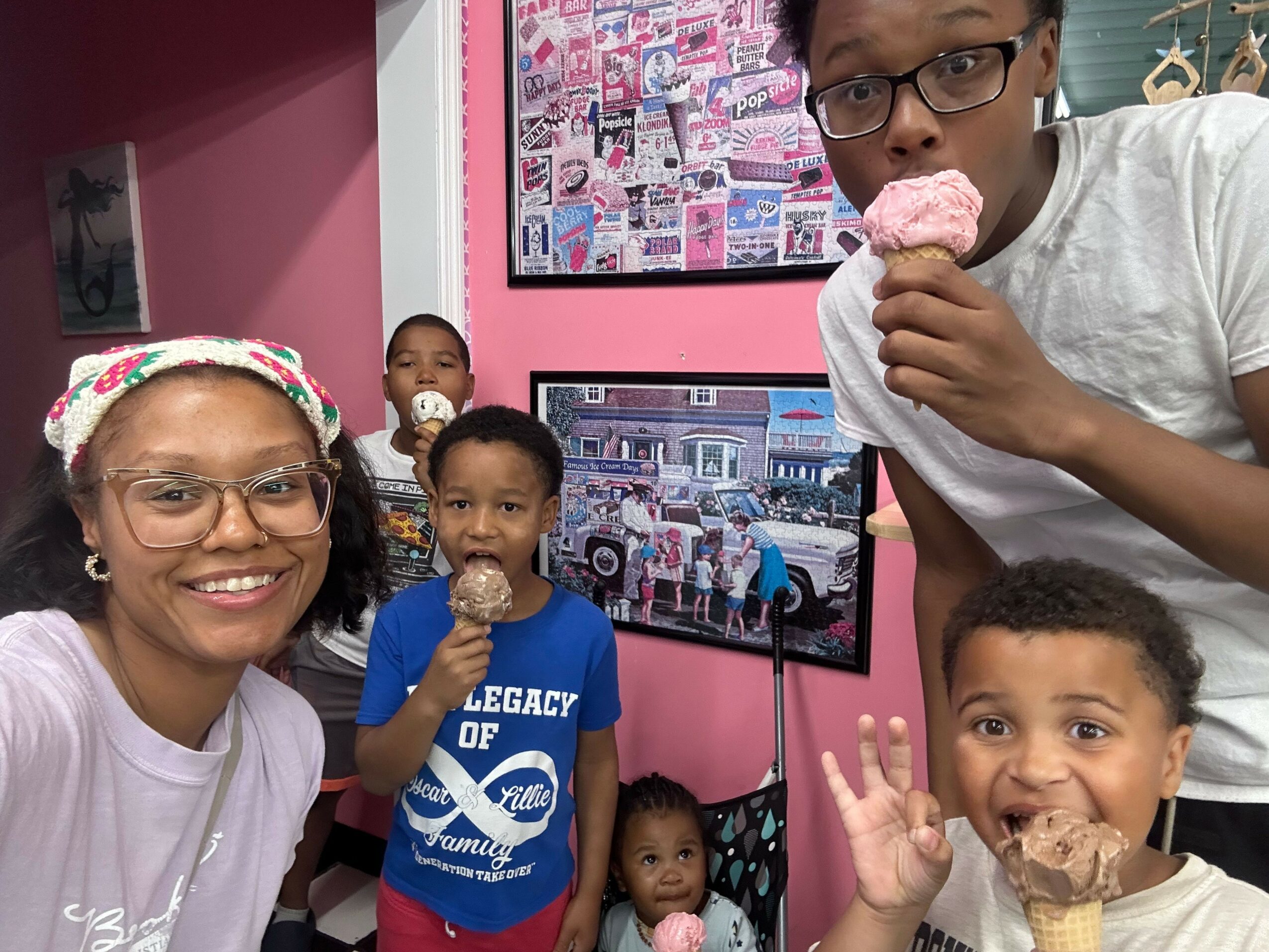 Group of children and an adult girl smiling and posing in a room with pink walls, with a computer screen displaying a colorful image.