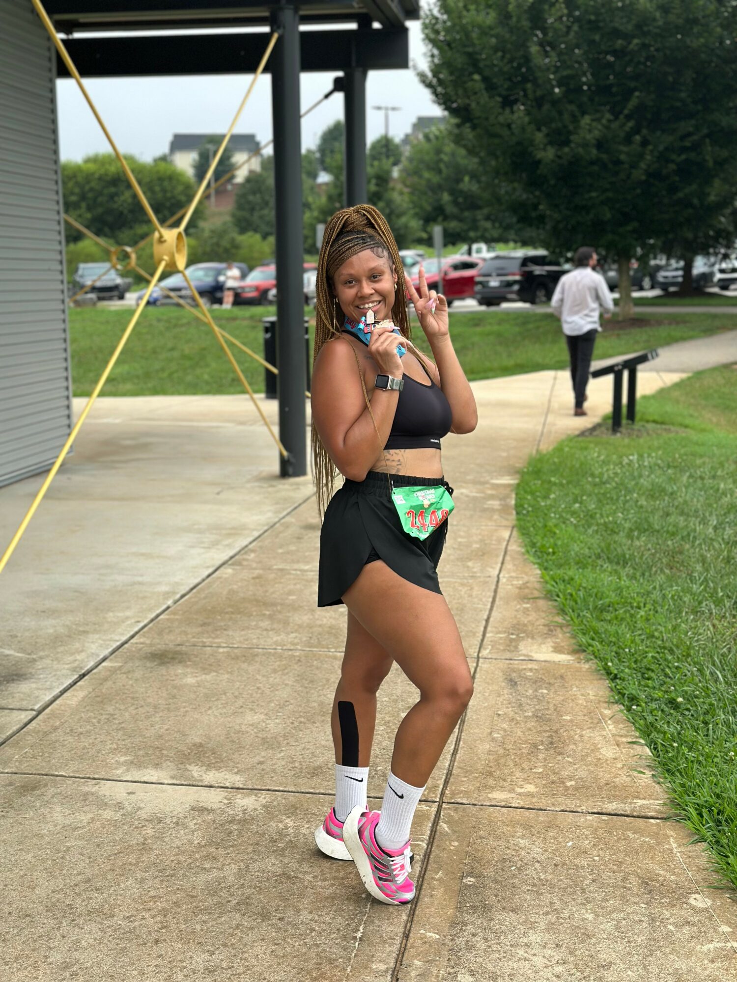 Young woman standing on sidewalk, smiling, wearing black sports bra, black shorts, pink and white sneakers, and a colorful fanny pack.