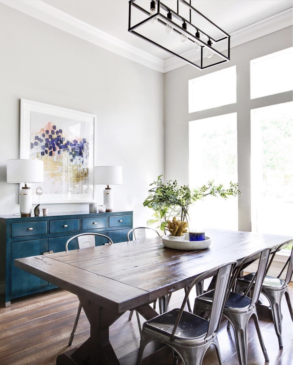 Dining room with wooden table, metal chairs, blue sideboard, large windows, and modern light fixture, bright and airy space.