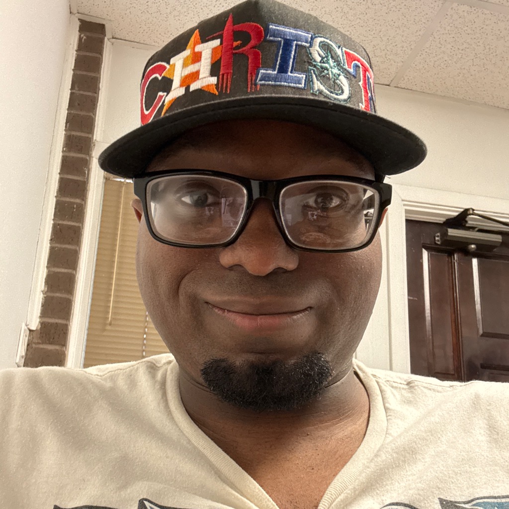Person wearing glasses and a colorful hat, smiling indoors with a brick wall and door in the background.
