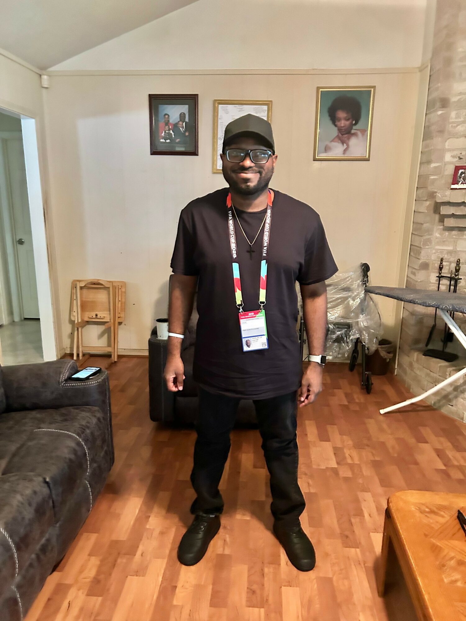 Man standing in living room, wearing black clothes, cap, sunglasses, and a lanyard, with framed pictures on wall behind him.