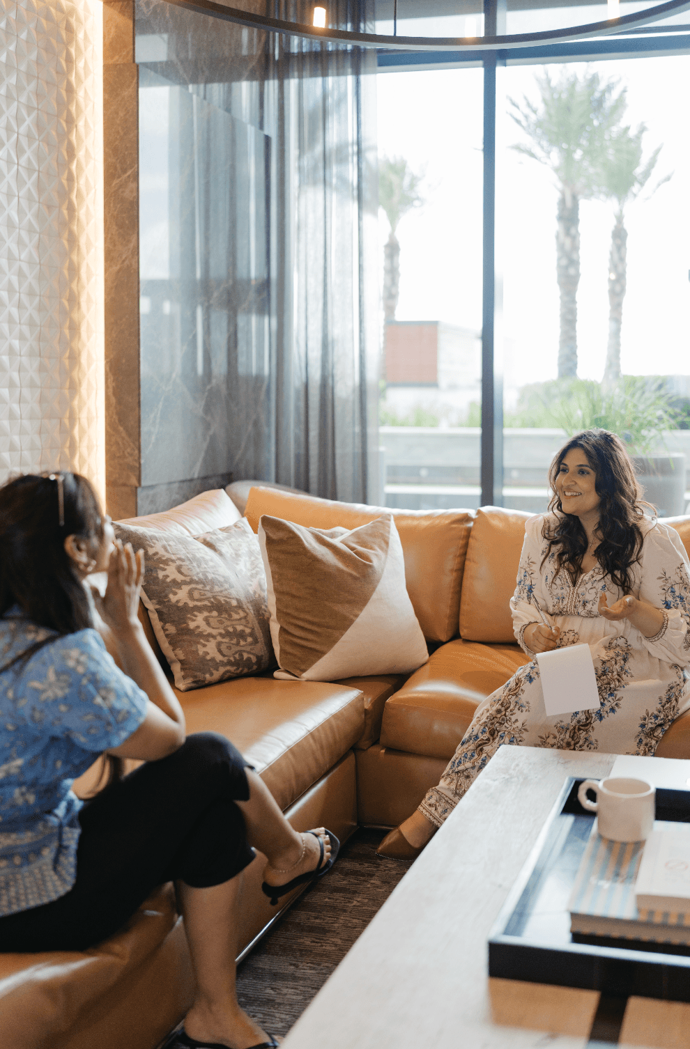 Two women sitting on a tan sofa in a bright room with large windows and palm trees outside, engaged in conversation.