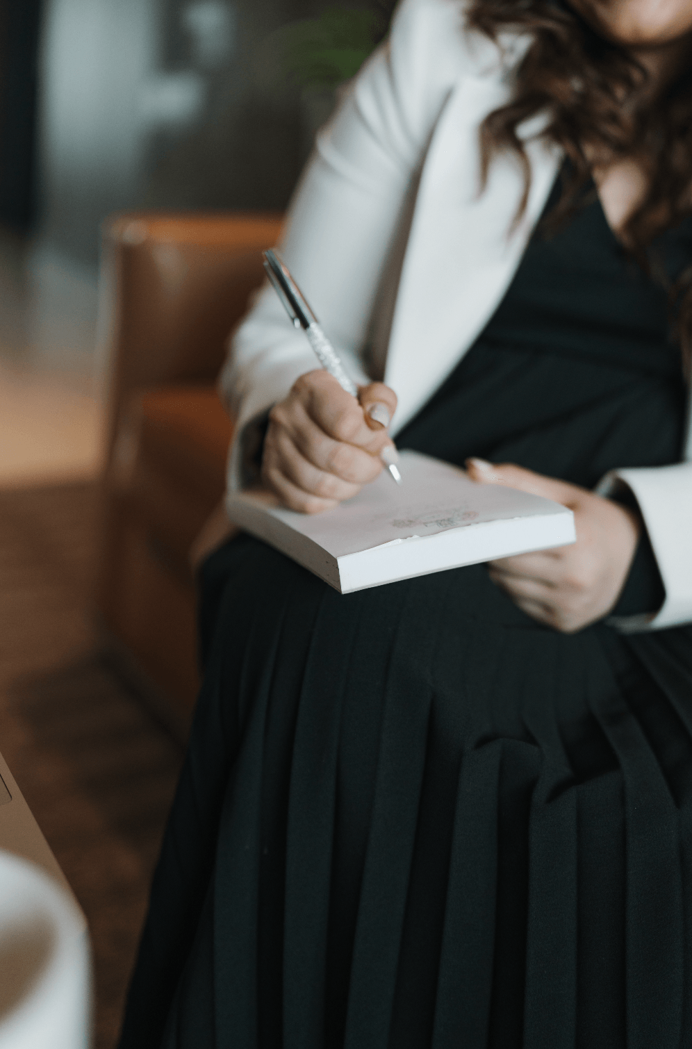 Person in white blazer writing in a notebook, sitting on a chair indoors, with blurred background.