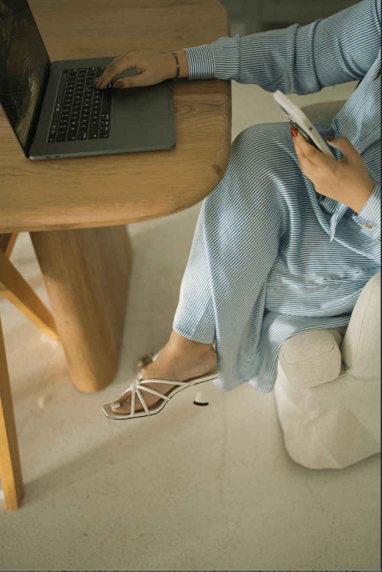 Person sitting at a wooden table using a laptop and holding a phone, wearing light-colored clothing and open-toe sandals.