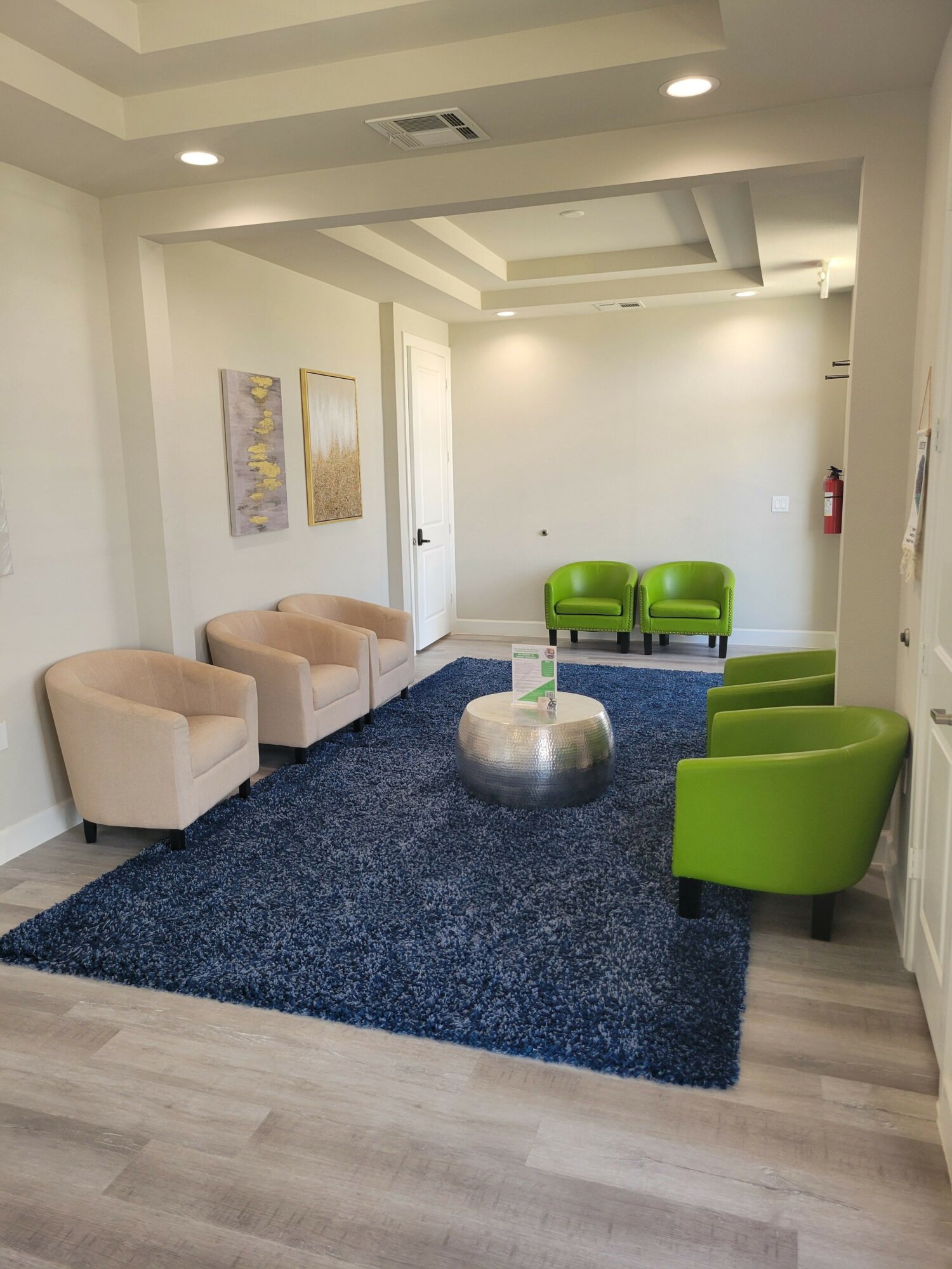 Modern waiting area with green chairs, beige ottomans, a blue rug, and artwork on white walls.