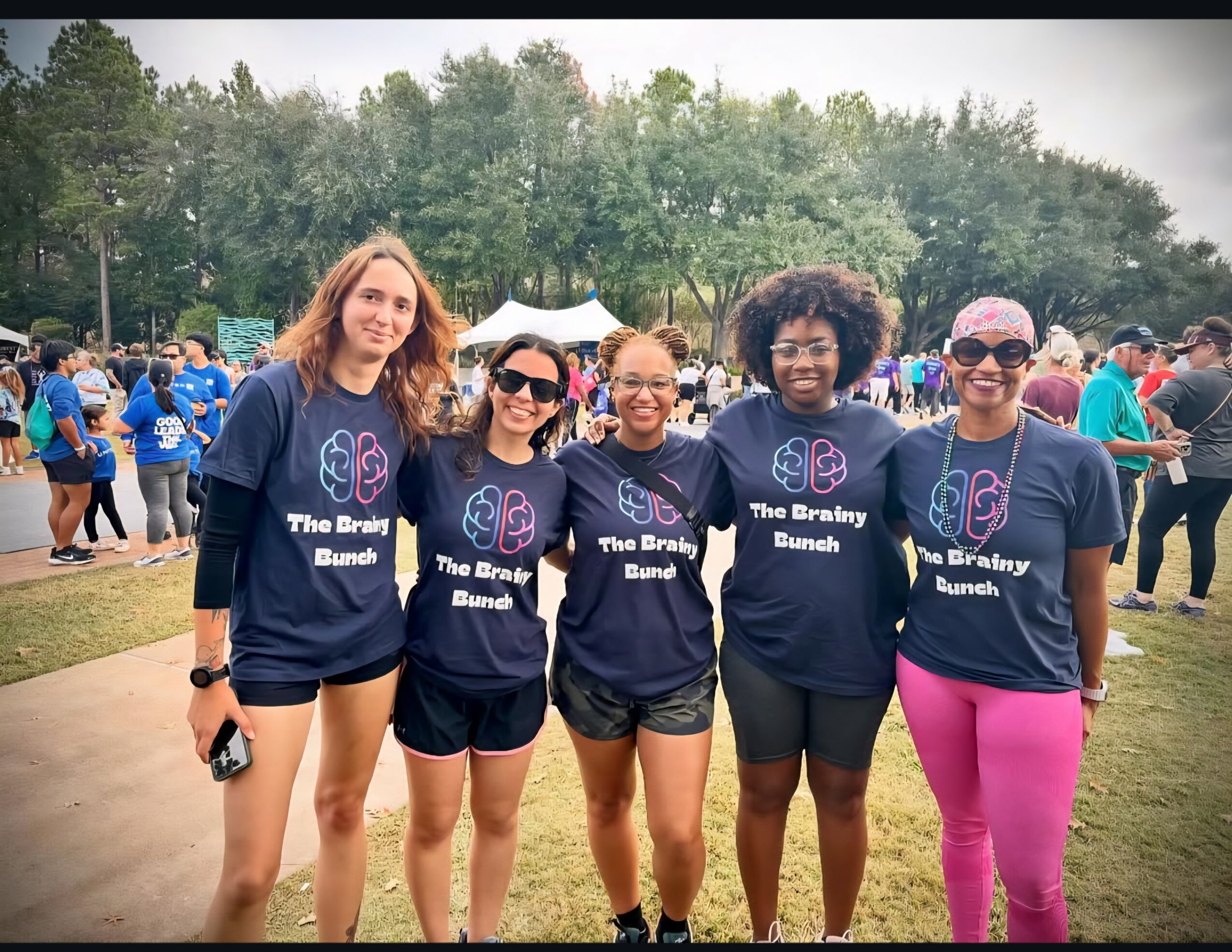 Five women standing outdoors in a park, wearing matching navy blue shirts, smiling, with trees and other people in the background.