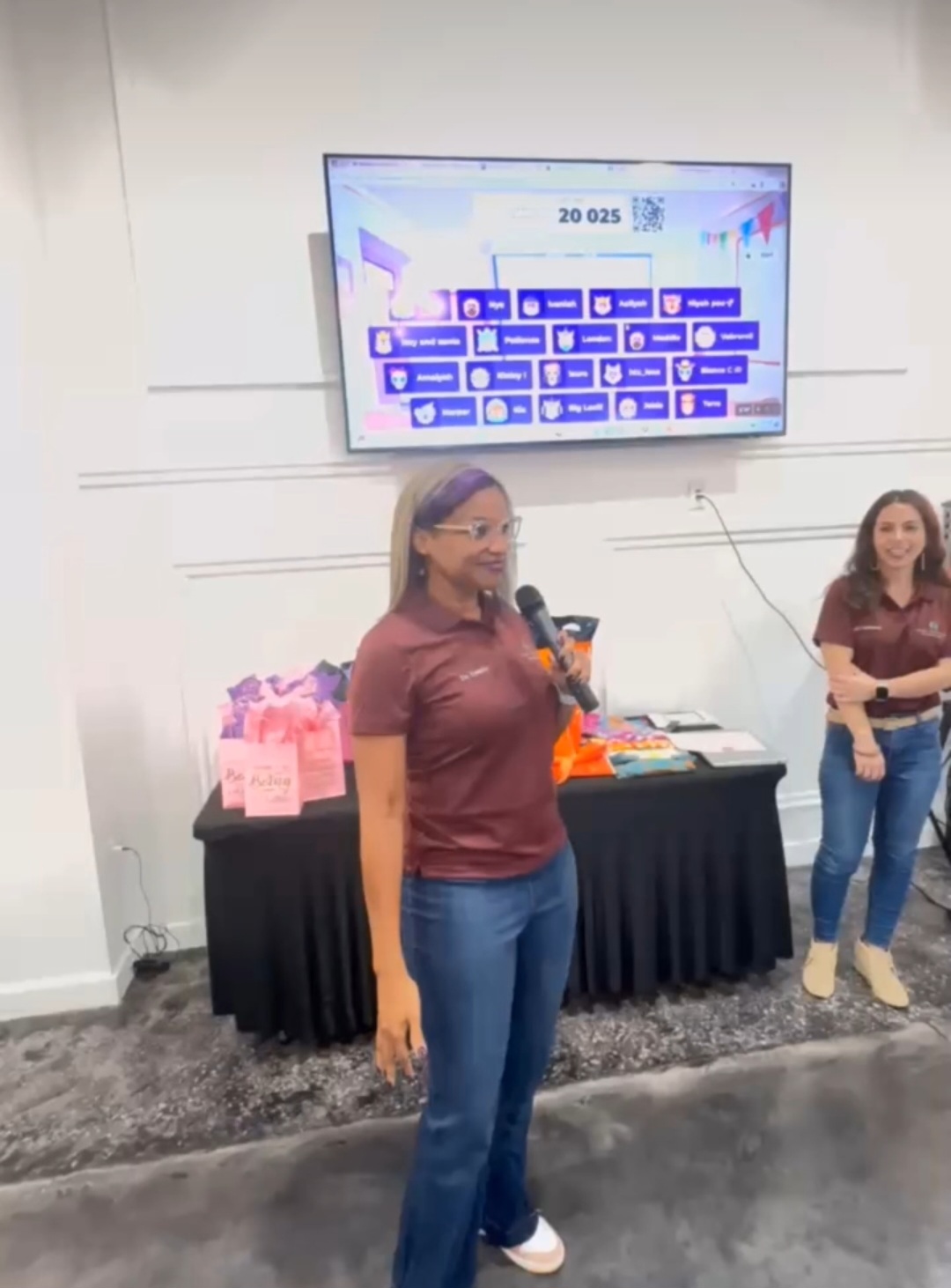 Two women standing in front of a table with pink items, one holding a microphone, in a room with a large screen above.
