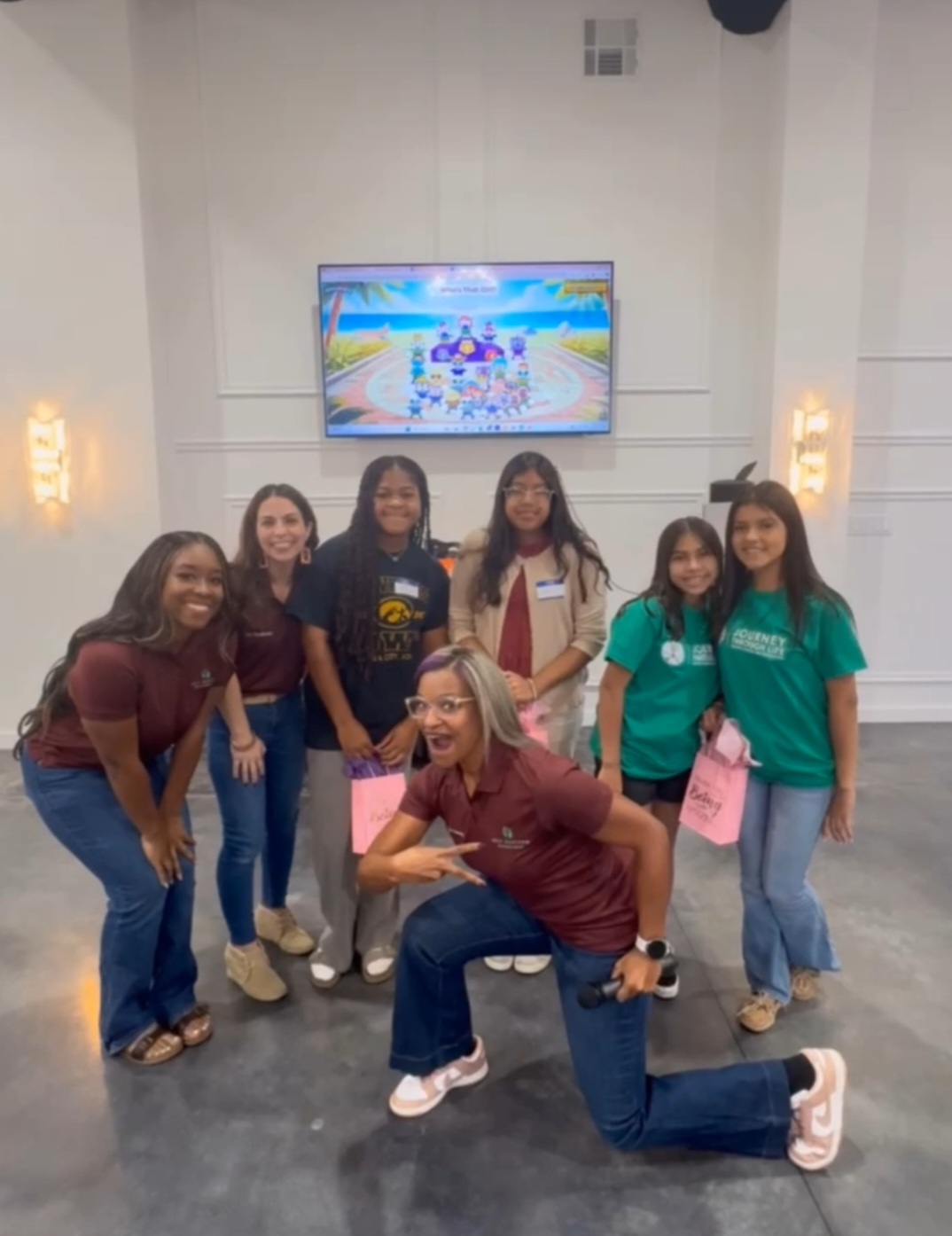 Group of seven women standing and one woman crouching in front, smiling in a room with a TV screen behind them.