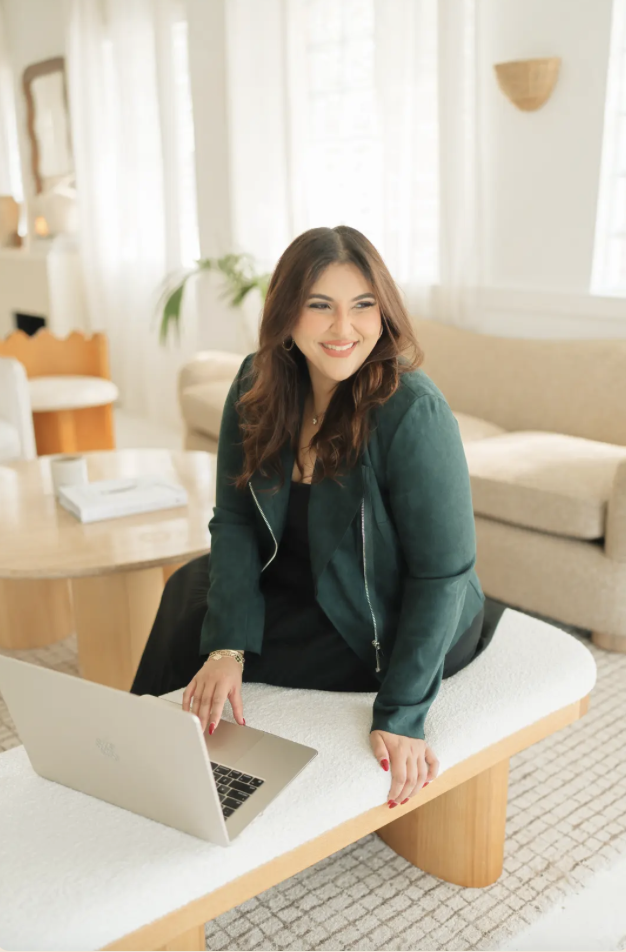 Young woman with long hair smiling at a laptop on a white table in a bright living room.