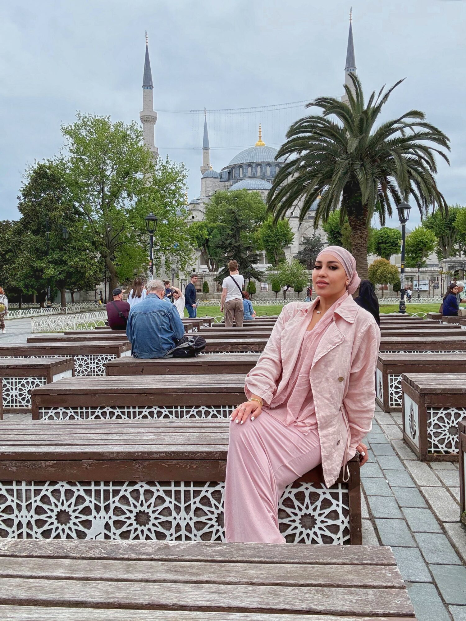 Woman in pink sitting on a bench in a park with trees, a mosque with minarets, and a palm tree in the background.