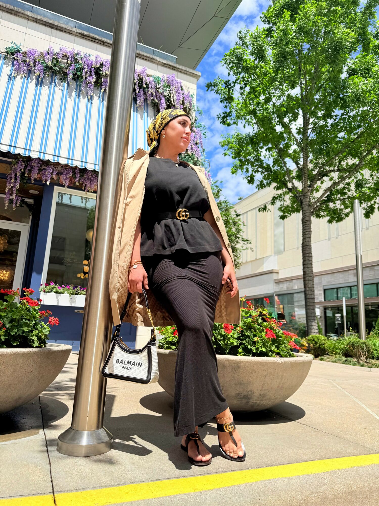 Woman standing outdoors near large planters and a tree, wearing black outfit and high heels, holding a shopping bag.