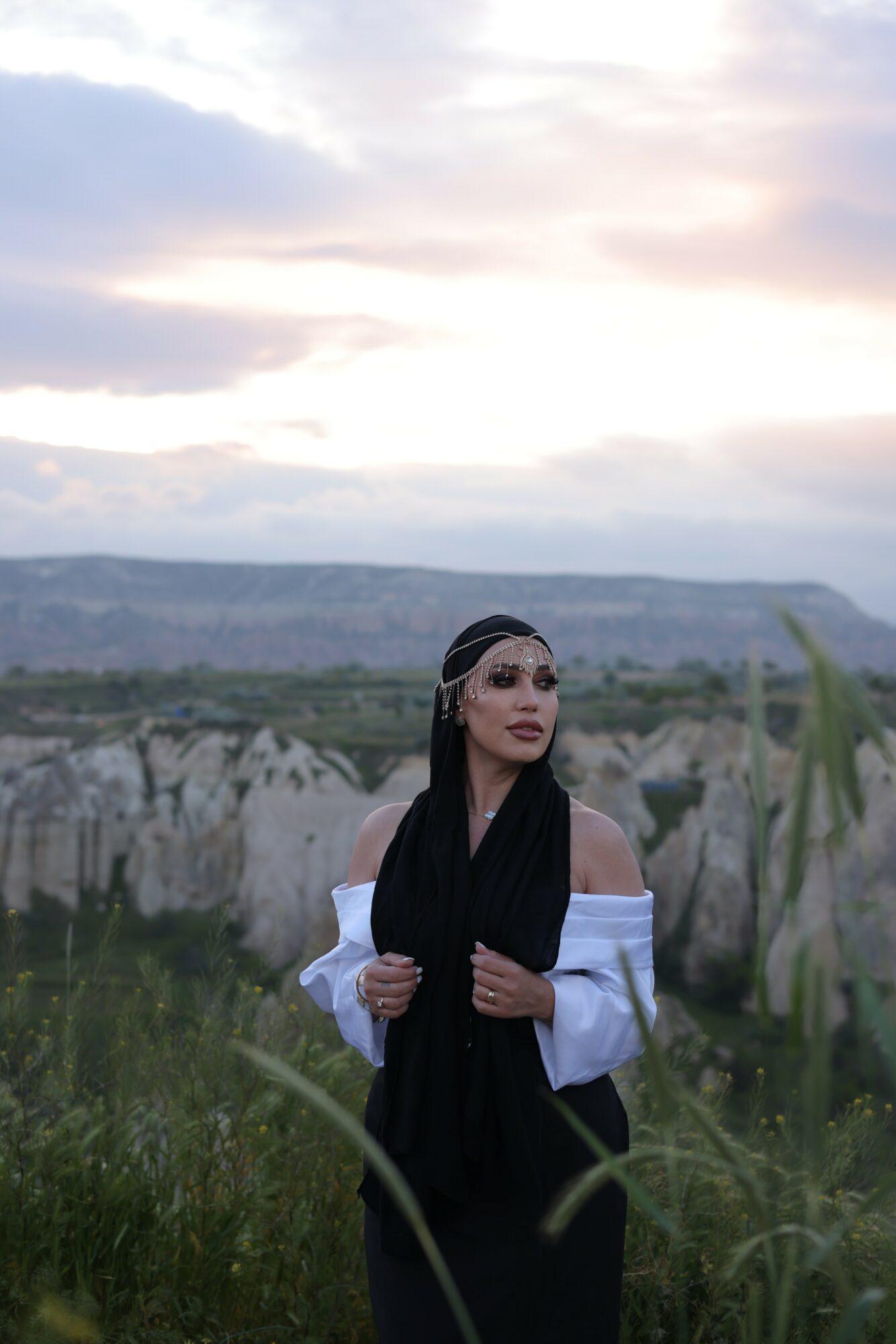 Woman with long dark hair and headband standing outdoors with cloudy sky and landscape background