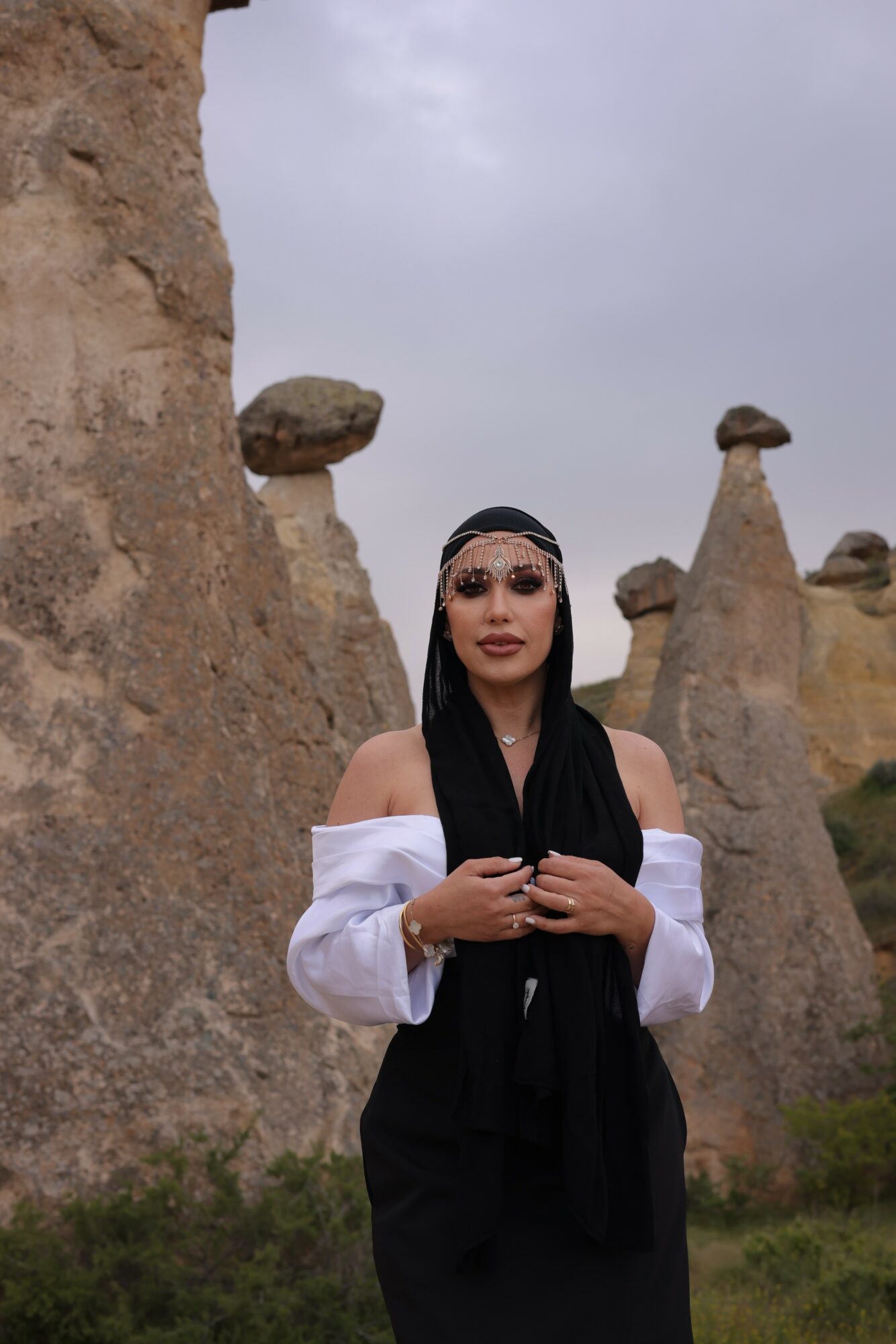 Woman standing outdoors with rock formations in the background, wearing a black headscarf and white off-shoulder top.