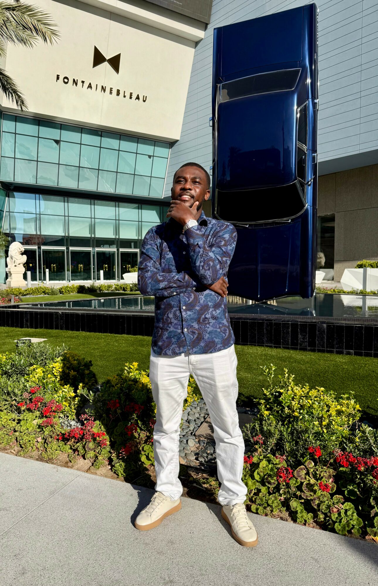 Young man standing outdoors in front of a modern building and a large blue sculpture, surrounded by colorful flowers.