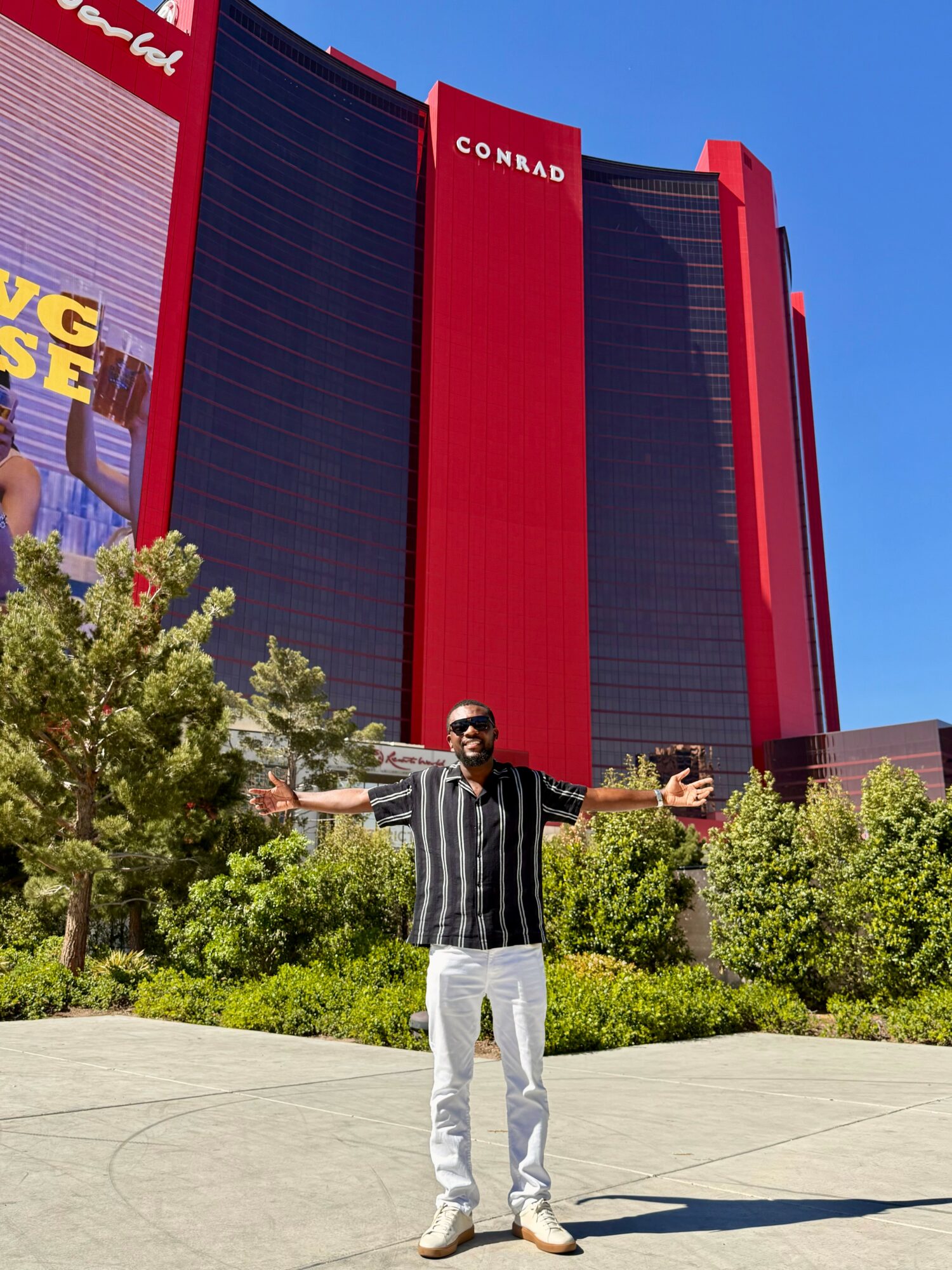 Person standing outdoors with arms outstretched in front of a large red and black building, trees and blue sky visible