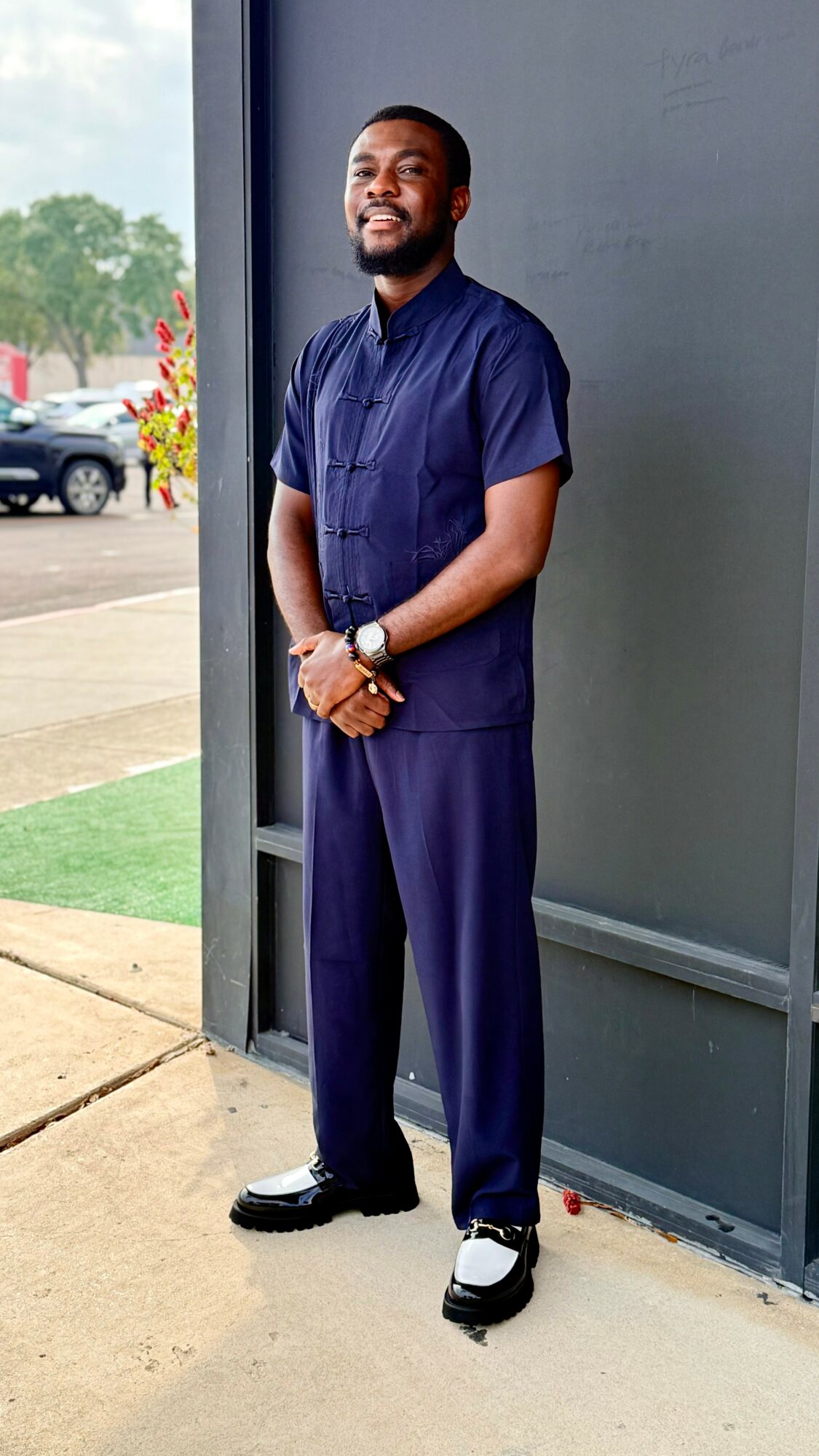 Man in navy scrubs standing outdoors near a building, smiling, with cars and trees in the background.
