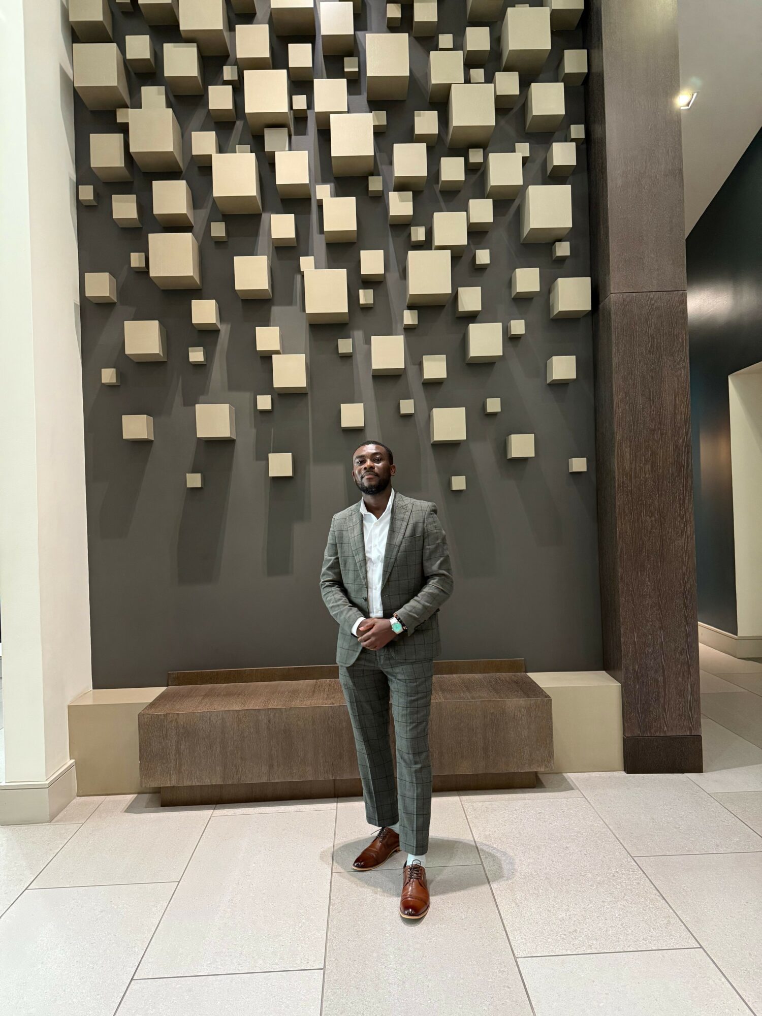 Person standing in front of a wall with 3D square decorations, wearing a gray coat and brown shoes.