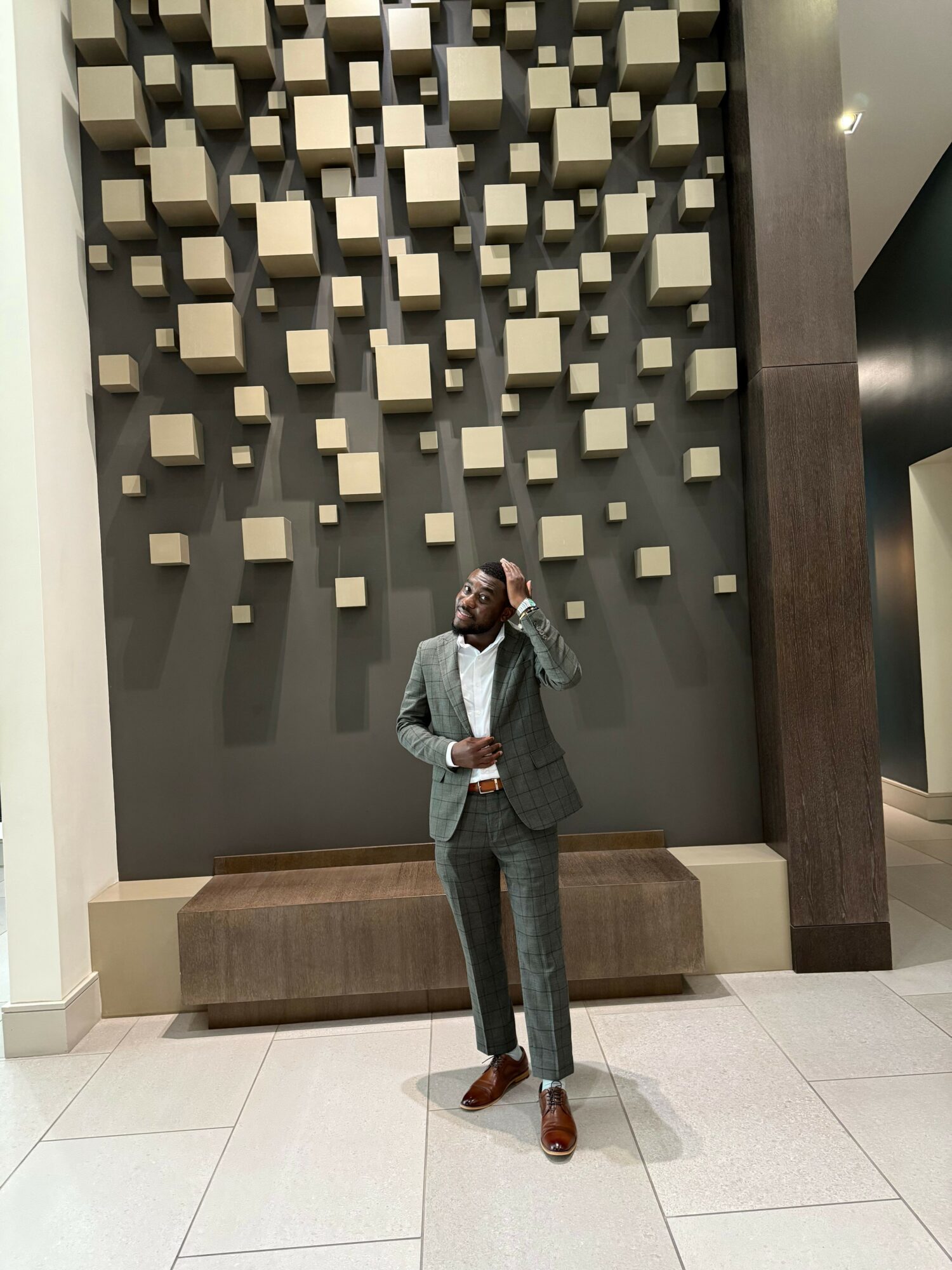 Person standing in front of a wall with 3D square decorations, wearing a suit and holding a bag, in an indoor setting.
