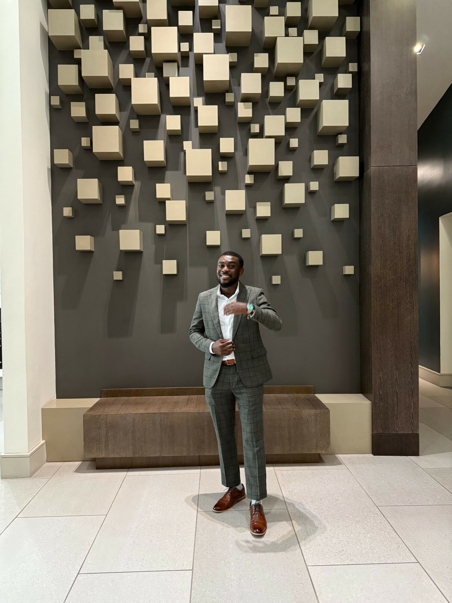 Person standing in front of a wall with 3D cube art, holding a bag, wearing a gray jacket and dark pants.