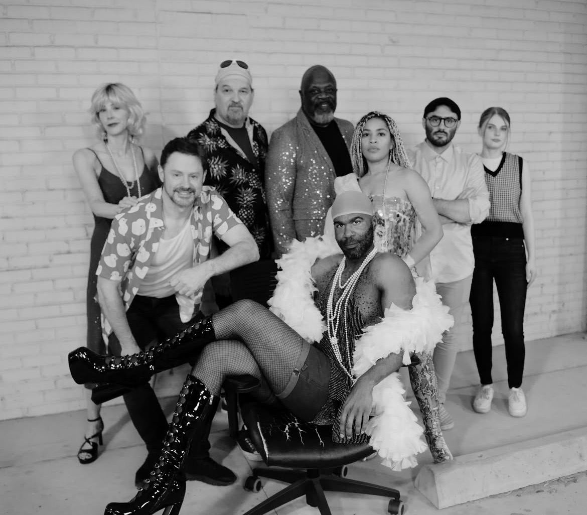 Group of nine diverse people posing against a brick wall, some standing and one sitting, in black and white.
