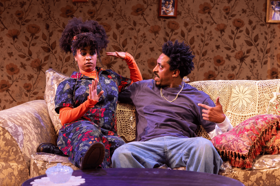 Two people sitting on a patterned sofa with a floral wallpaper background, engaging in conversation, one with curly hair and colorful clothing, the other with short curly hair and casual attire.