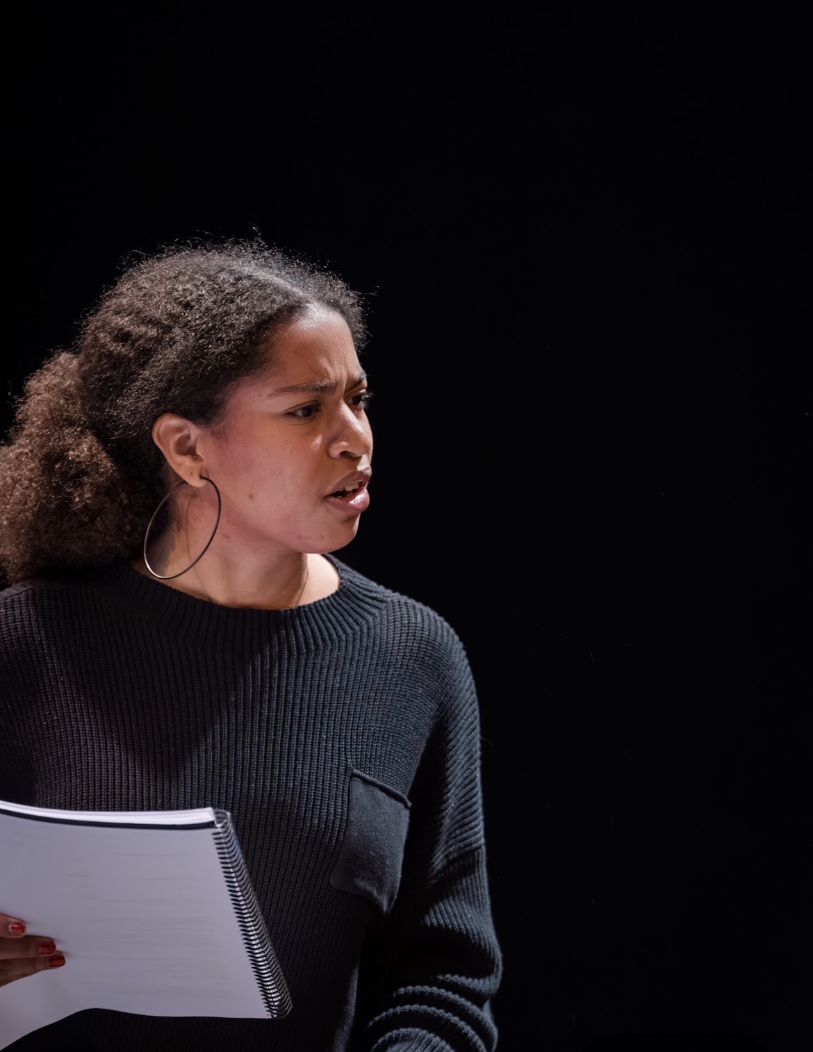 Woman with curly hair holding a notebook, wearing a dark sweater, against a black background.