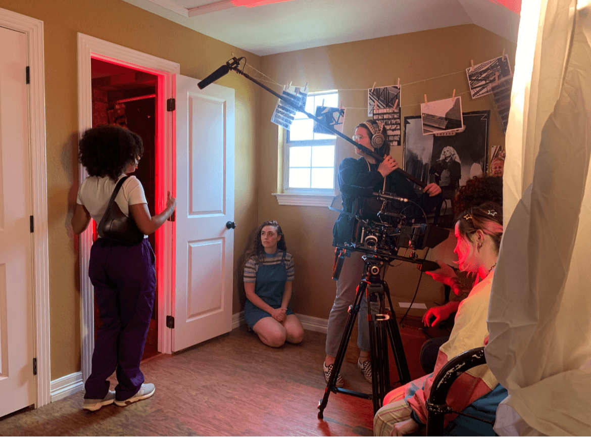 Group of people in a room with a camera, some sitting and others standing, with photos on the wall.