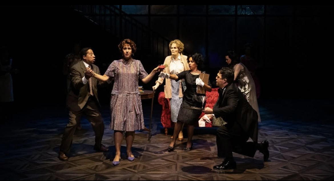 Group of seven people on stage, some holding hands, in a theatrical scene with dark background.