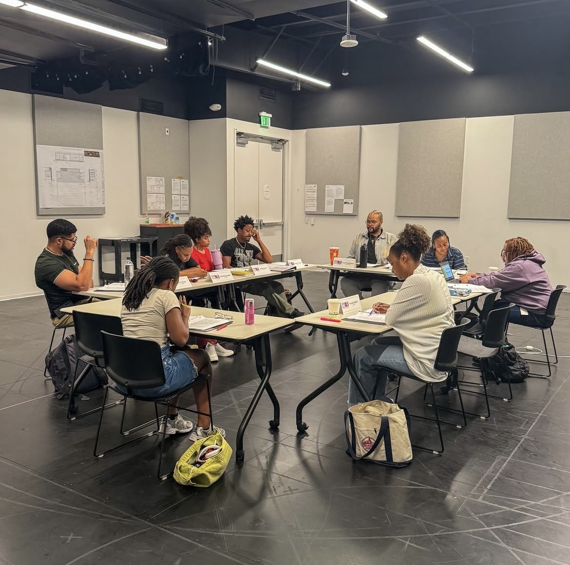 People sitting around tables in a classroom or meeting room, engaged in discussion or note-taking.