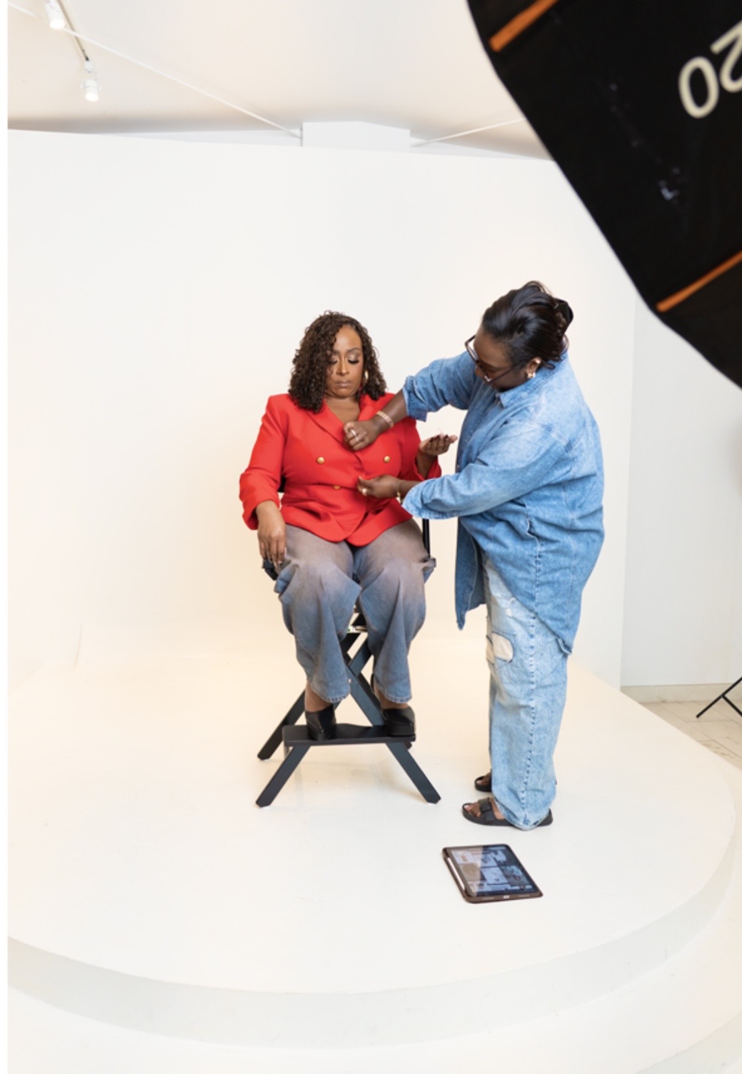 A woman in a red jacket sits on a stool while a person in a blue outfit assists her with a medical procedure in a studio setting.