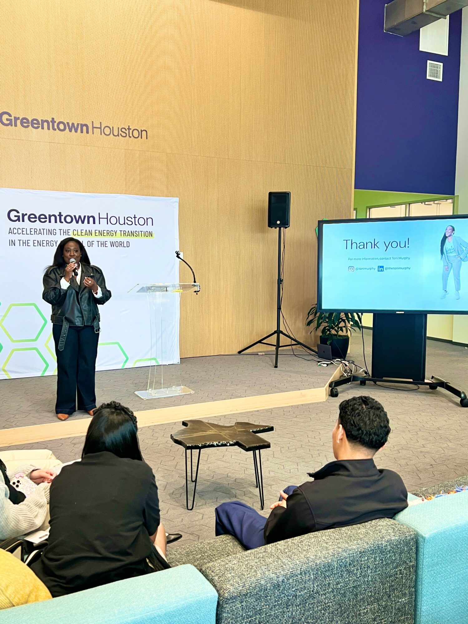 Person speaking on stage at Greentown Houston event, audience seated, large screen displays message, background features blue and green colors.