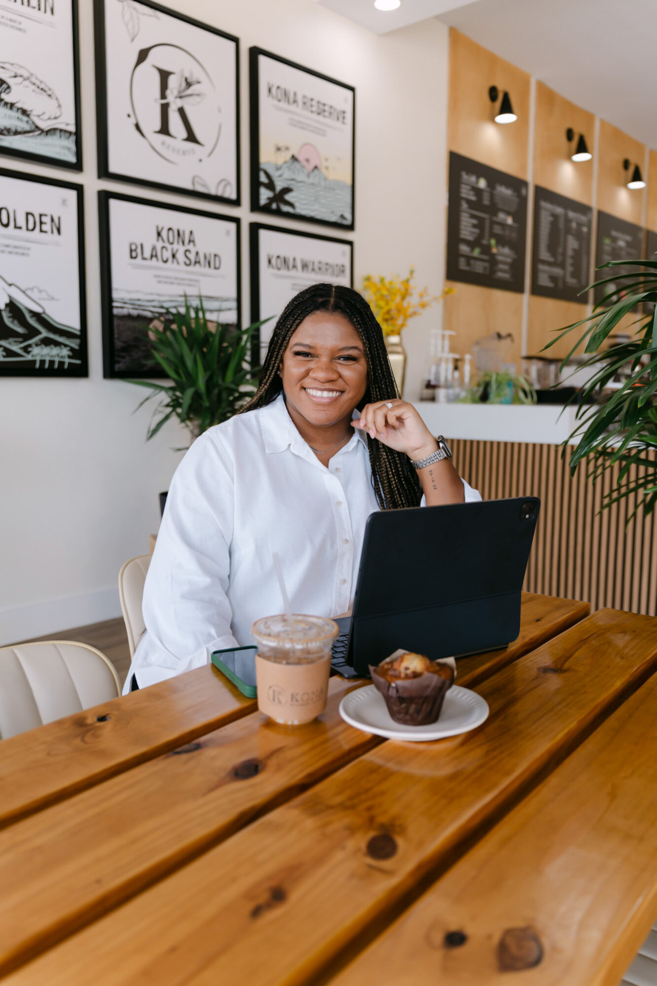 Smiling woman sitting at a wooden table with a laptop, drink, and dessert in a cafe with framed posters and plants.