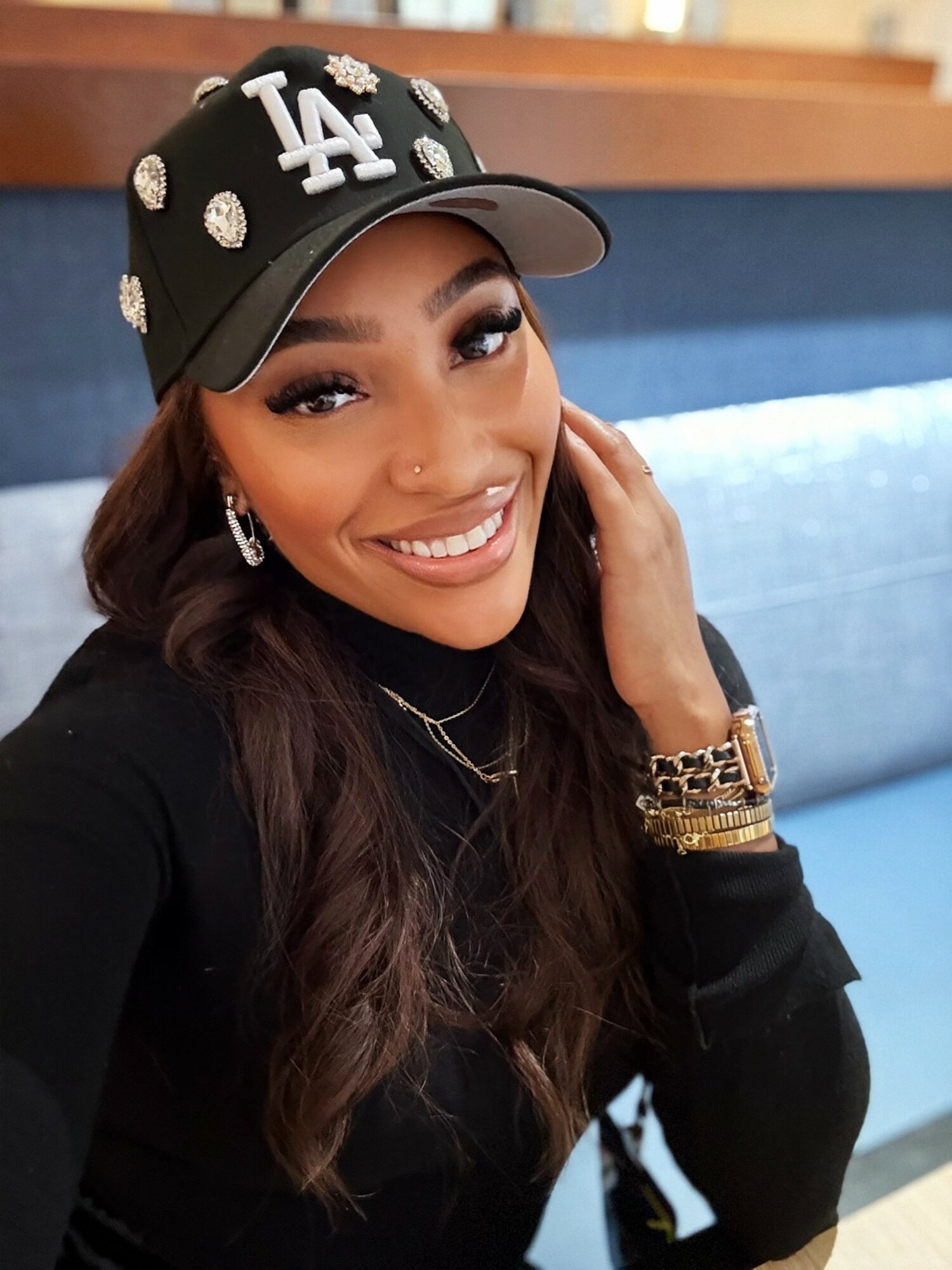 Smiling woman wearing a black LA cap, earrings, and gold jewelry, sitting indoors with a blue background.