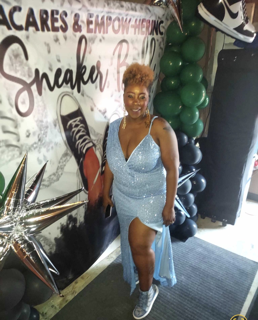 Smiling woman with short curly hair in a light blue dress and sneakers, standing in front of a decorated backdrop with balloons.