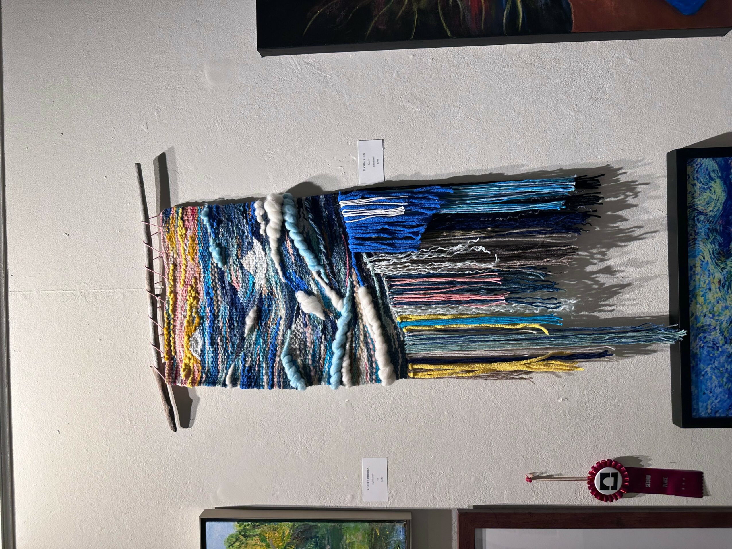 Colorful woven textile art piece hanging on a wall with a small label below it, and a television screen partially visible on the right.