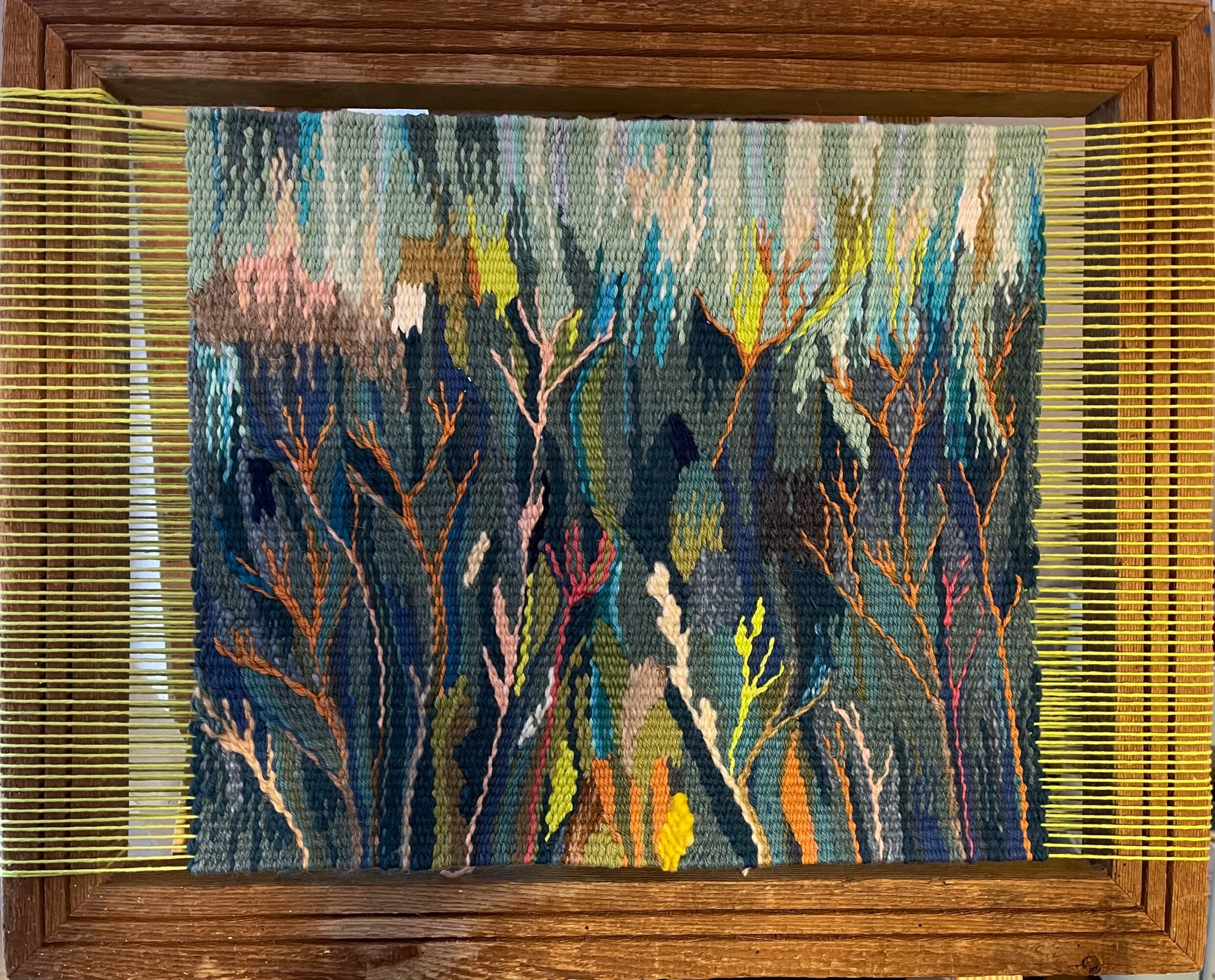 Colorful abstract textile with plant-like shapes, framed by yellow slats, on a wooden background.