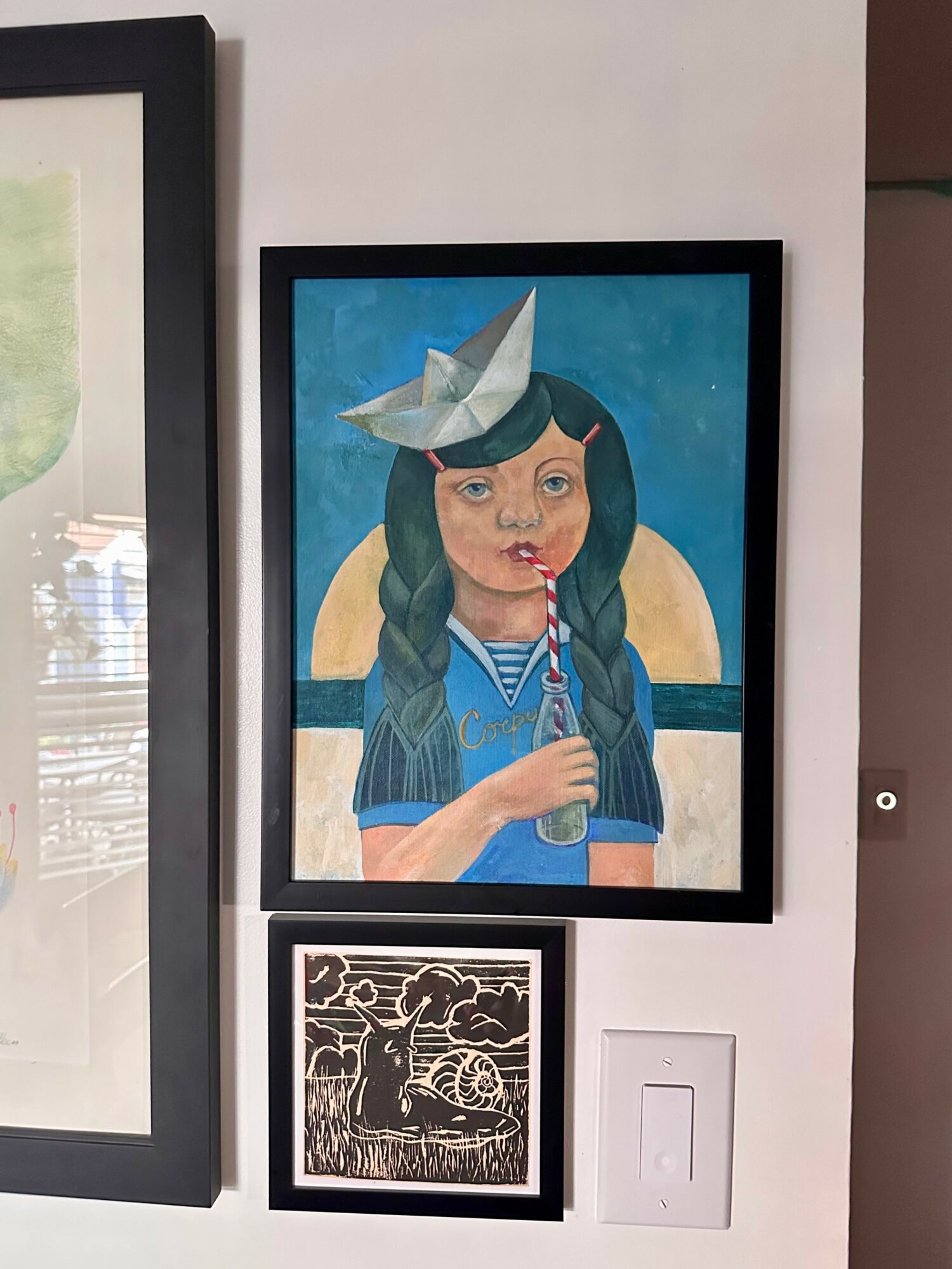 Framed painting of a girl with long dark hair, wearing a blue shirt, holding a bottle, with a paper hat on her head, against a blue background with a yellow circle. Smaller black and white artwork below, and a light switch to the right.