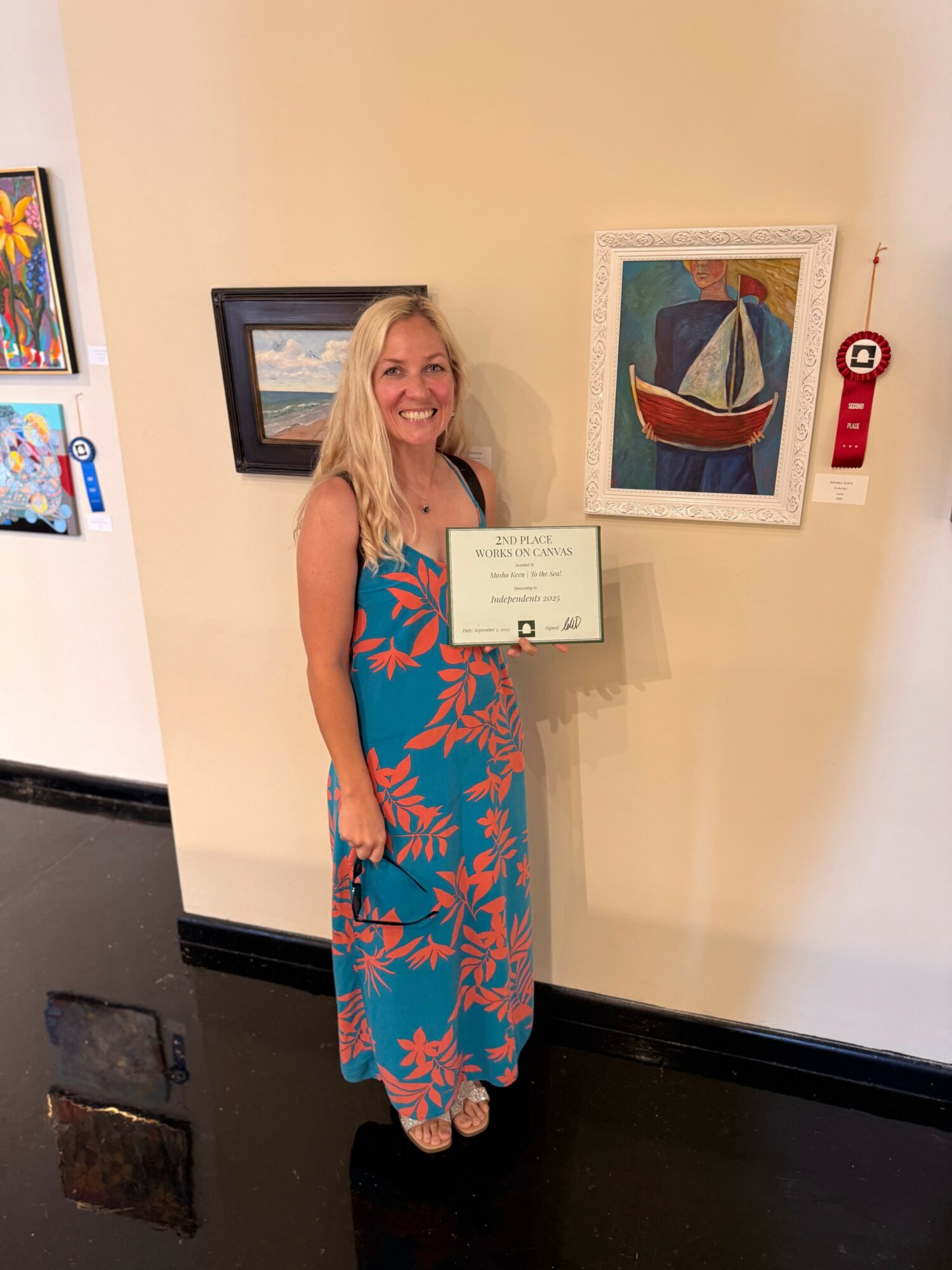 Woman with long blonde hair in a colorful dress standing next to framed artwork on a beige wall.