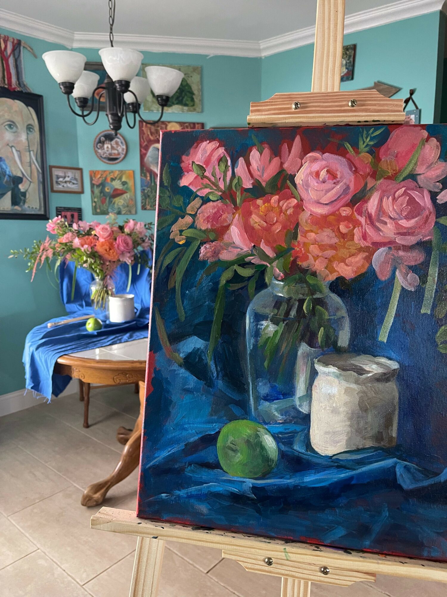 Painting of pink flowers in a vase on a blue background, with a white object and green leaf, on an easel.