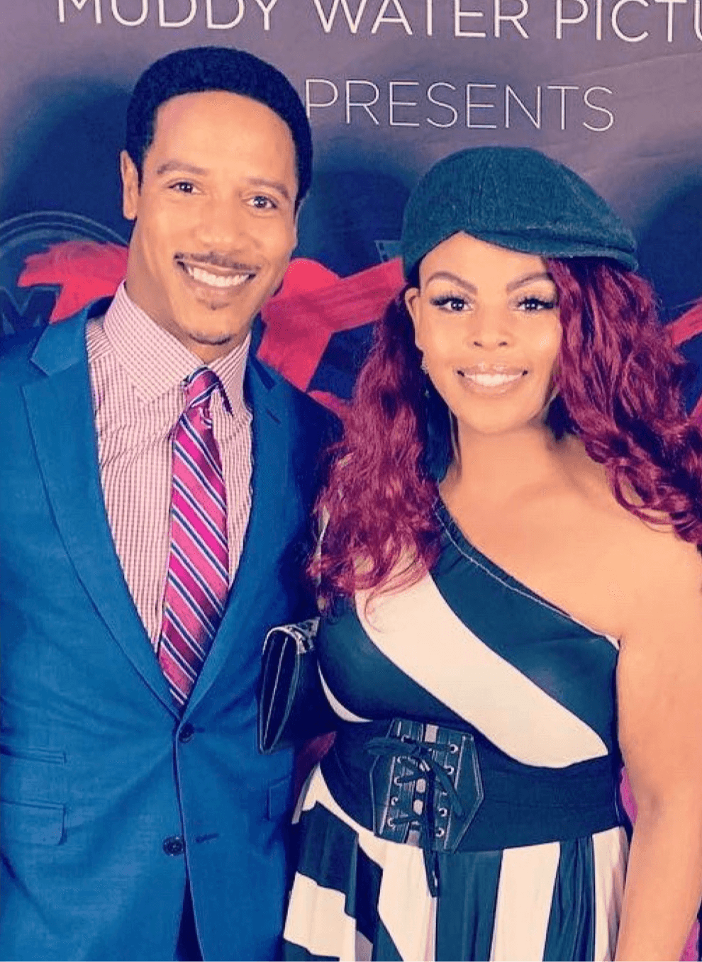 Two people smiling, one man in a blue suit and striped tie, woman with curly hair in a striped dress and beret.