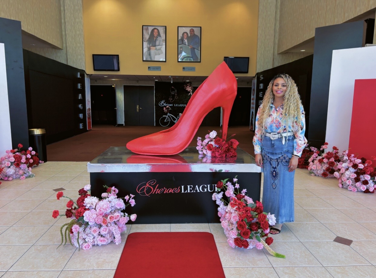 Large red high-heeled shoe display with flowers, woman in blue dress, and posters in background.