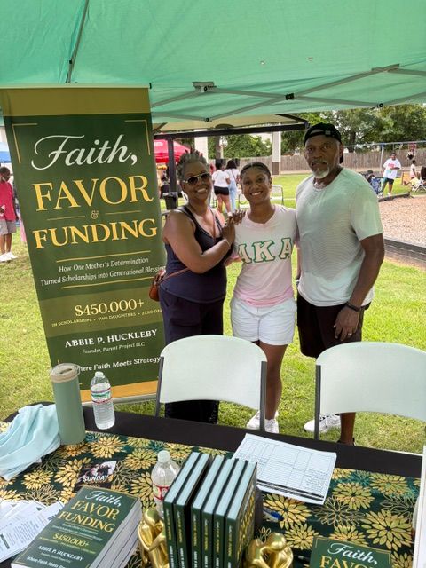 Three people standing behind a table with books and a sign at an outdoor event, green canopy overhead.