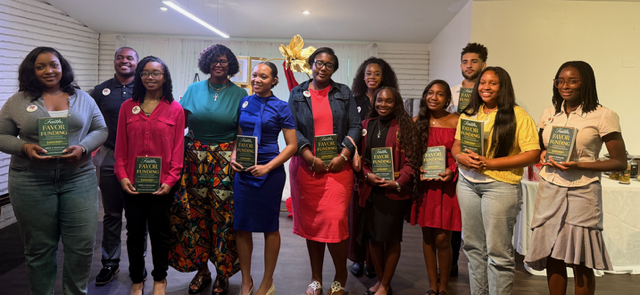 Group of people standing indoors, holding books, smiling, with a decorative plant in the background.
