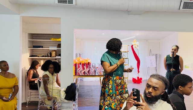 Group of people in a room with a woman in colorful clothing speaking, others seated or standing, some using phones.